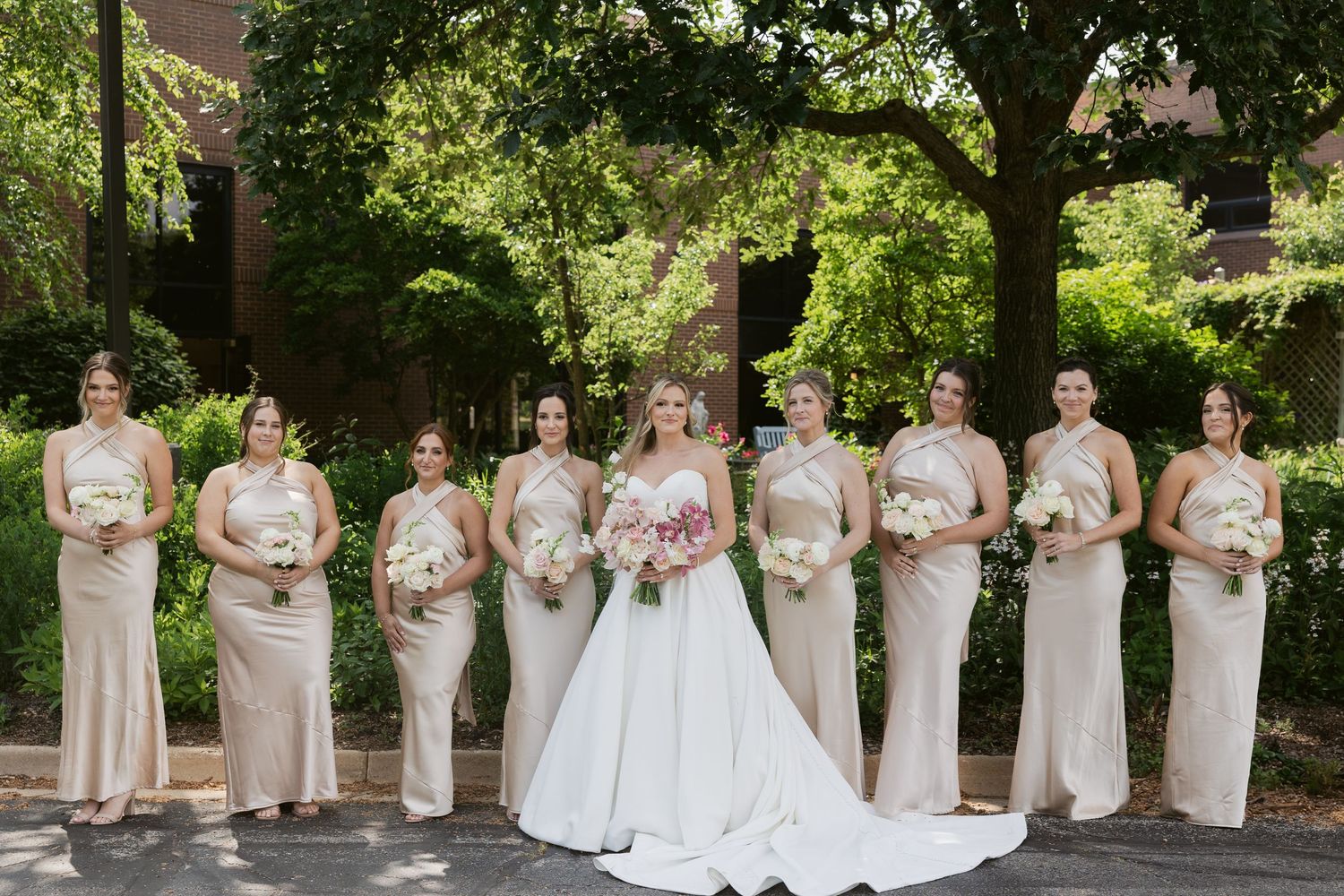 Wedding party in champagne dresses posed outdoors with bride in white gown under lush green trees.