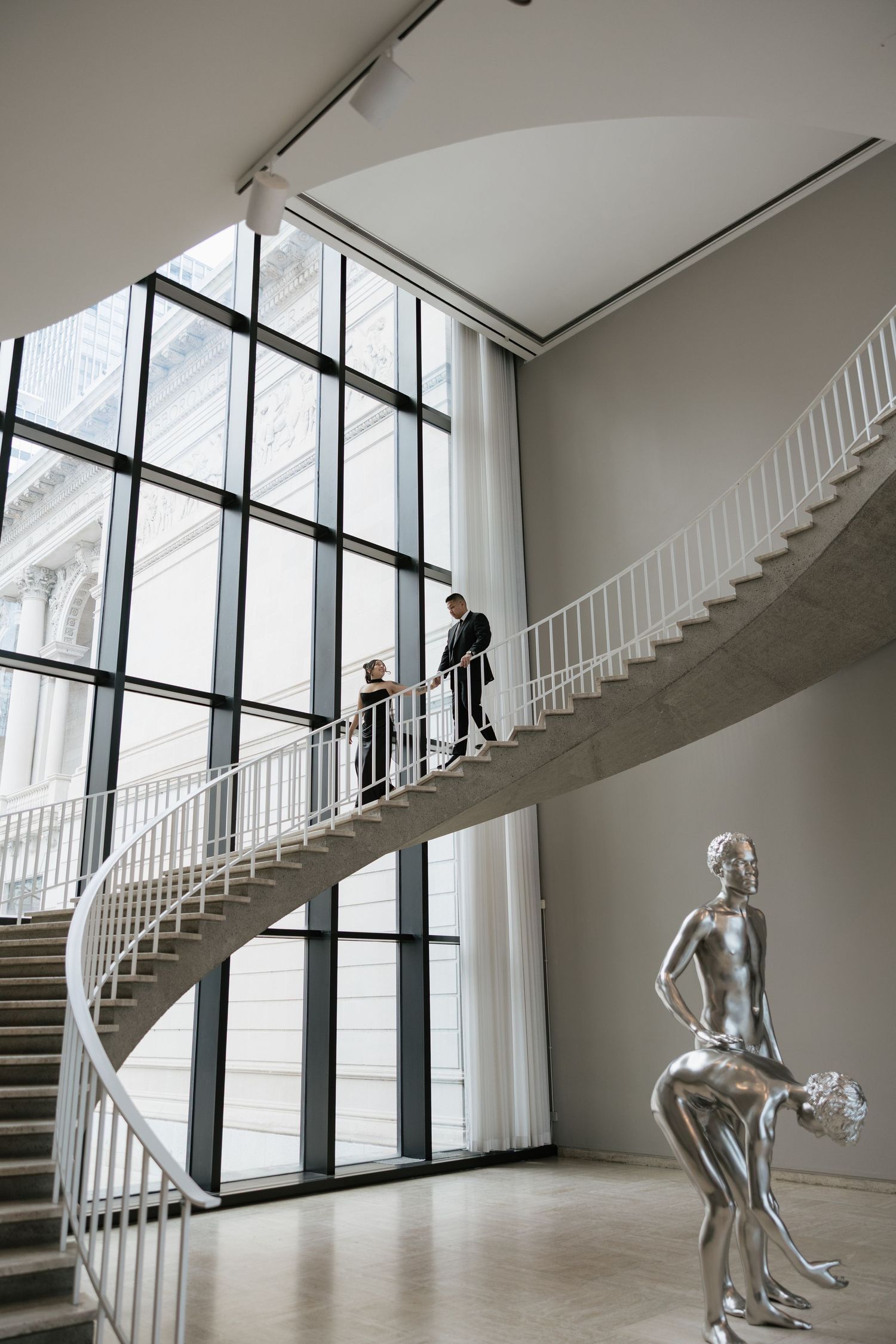 A dramatic spiral staircase in a modern museum with floor-to-ceiling windows and a silver sculpture in the foreground.