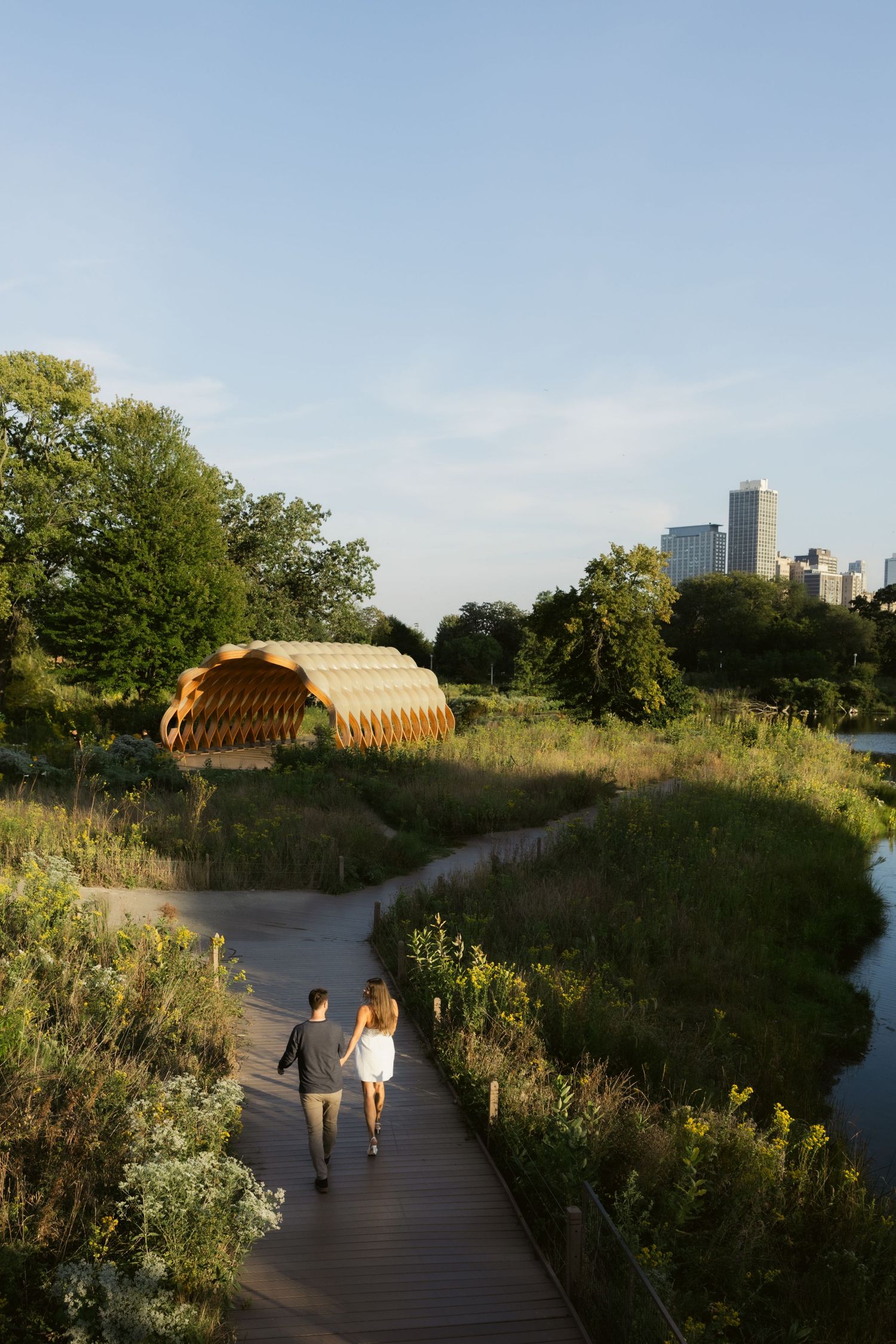 A wooden boardwalk path winds through natural vegetation with city buildings visible in the background.