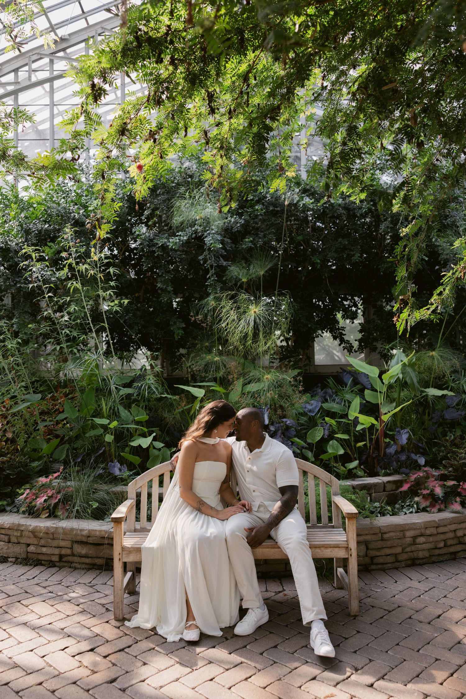 A couple in white outfits sits together on a wooden bench inside a greenhouse filled with tropical plants.