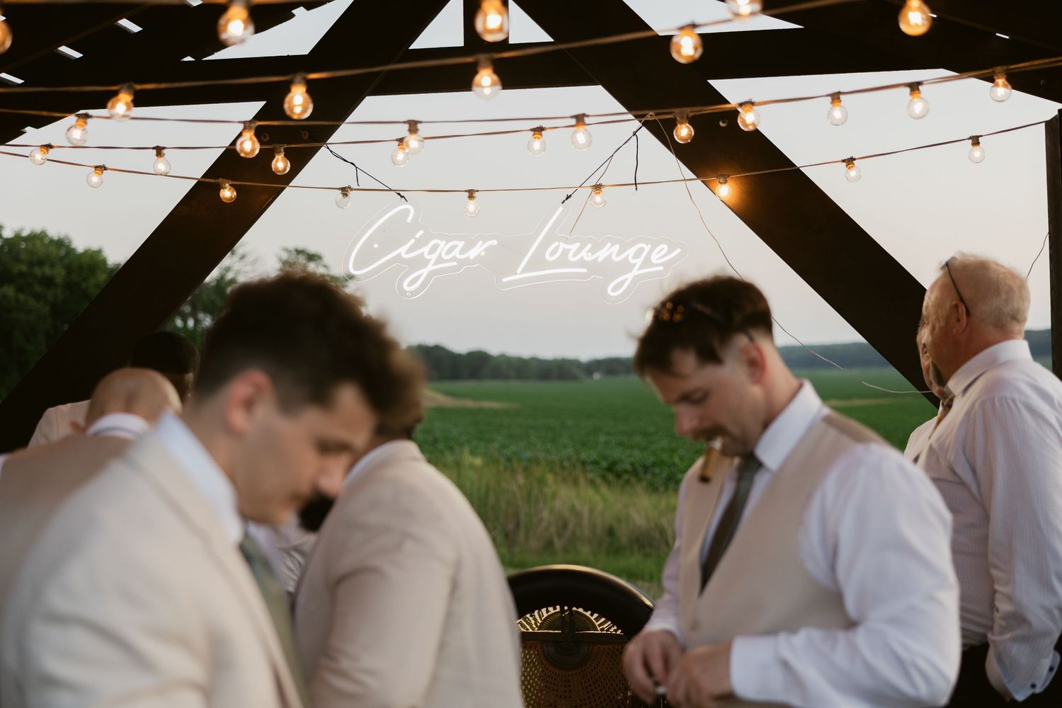 People gather for a rustic outdoor wedding reception under string lights and white tents at dusk.