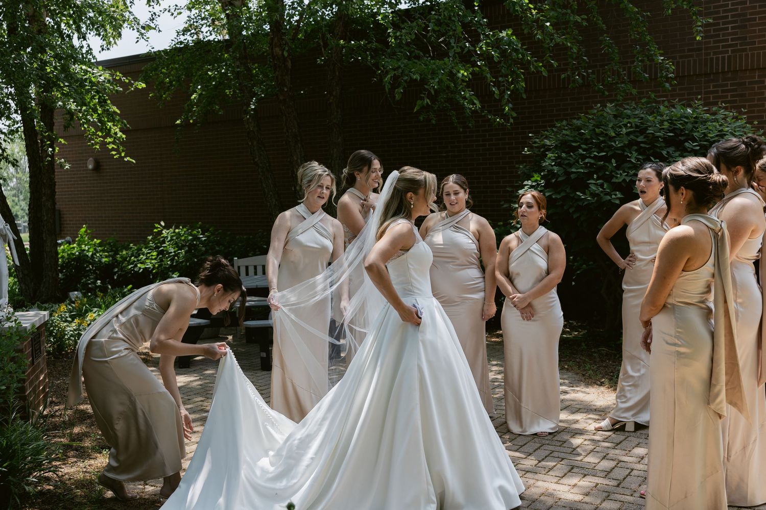 Wedding party in neutral dresses assisting with the bride's gown on a sunny outdoor pathway.