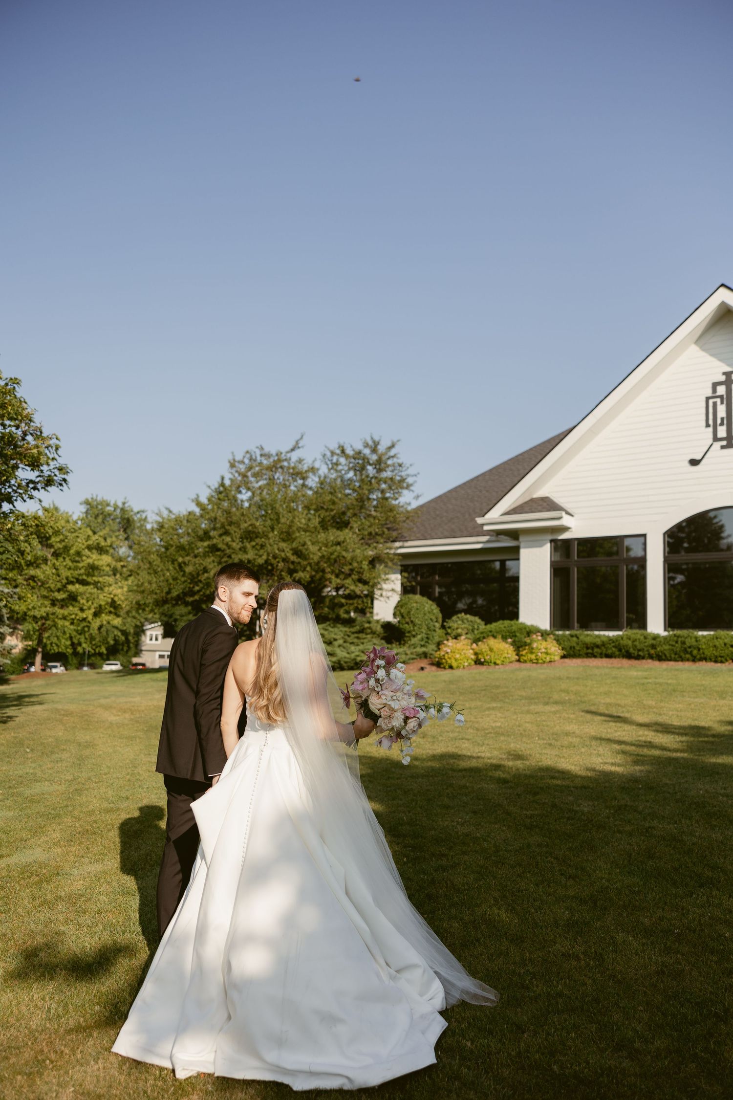 Newlywed couple dances on a sunny lawn with white buildings in the background.