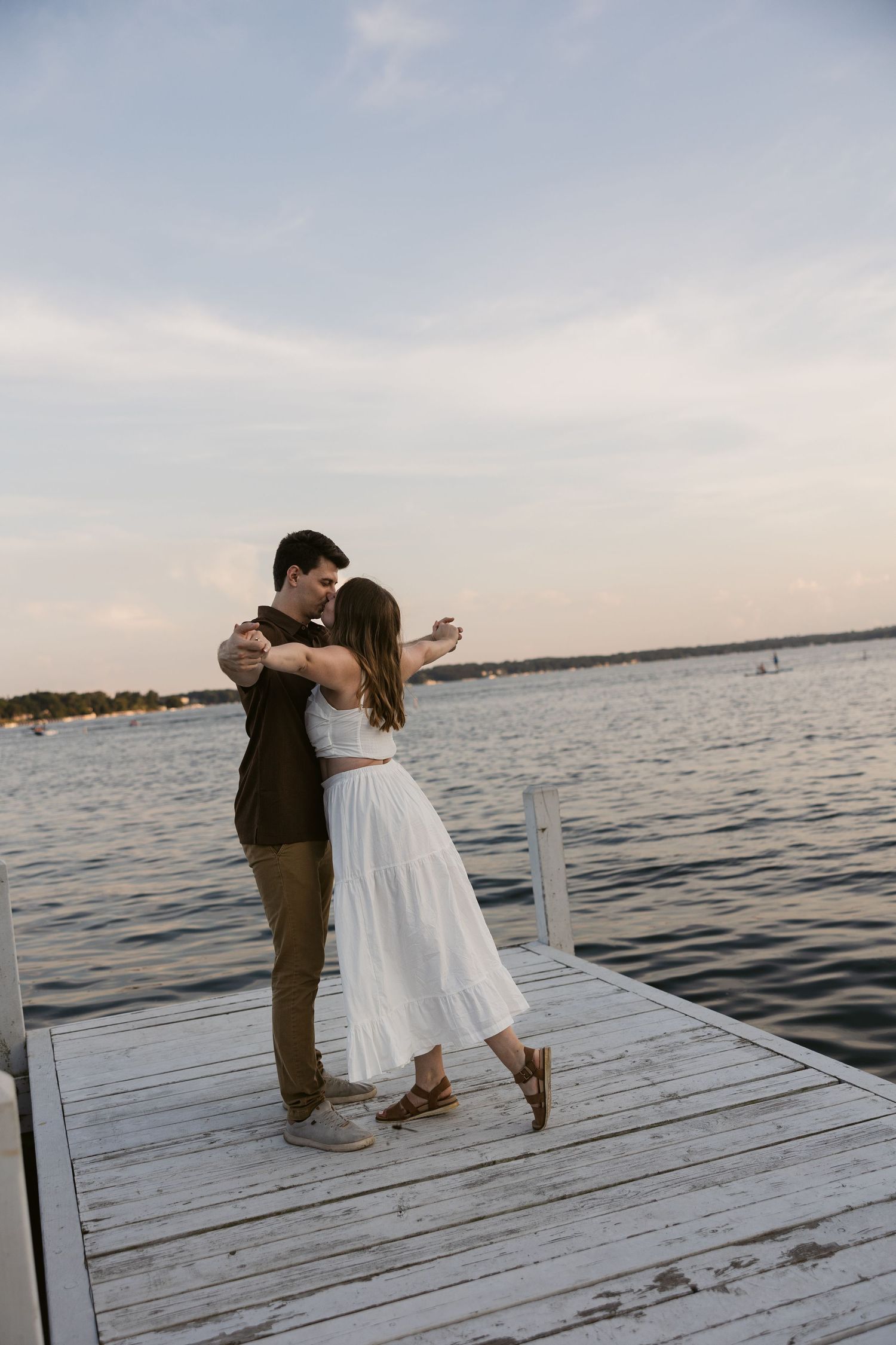 A romantic couple embraces on a white dock overlooking a serene lake at sunset.