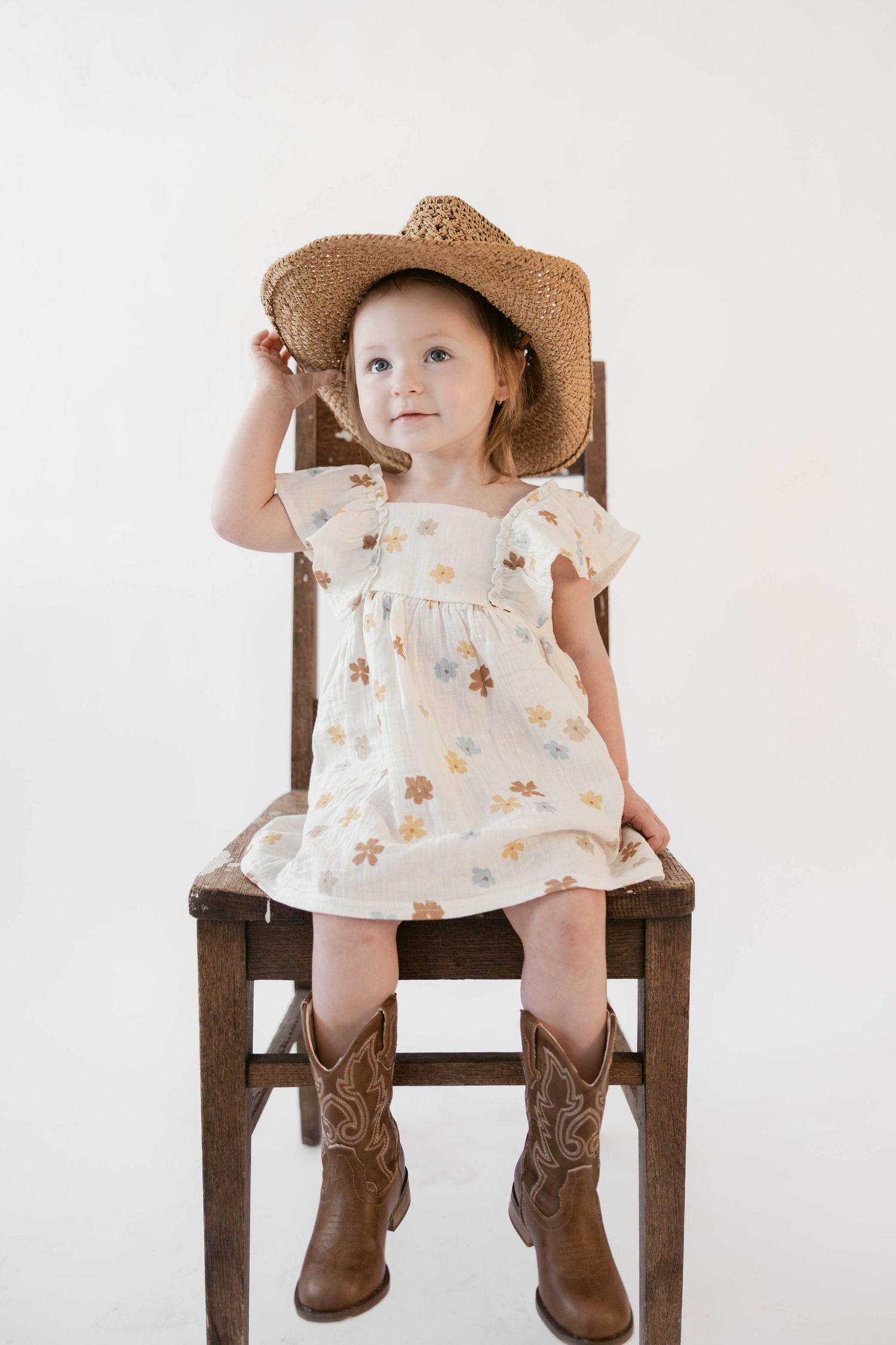 A toddler wearing a floral dress and cowboy boots sits on a wooden chair while wearing a straw hat.