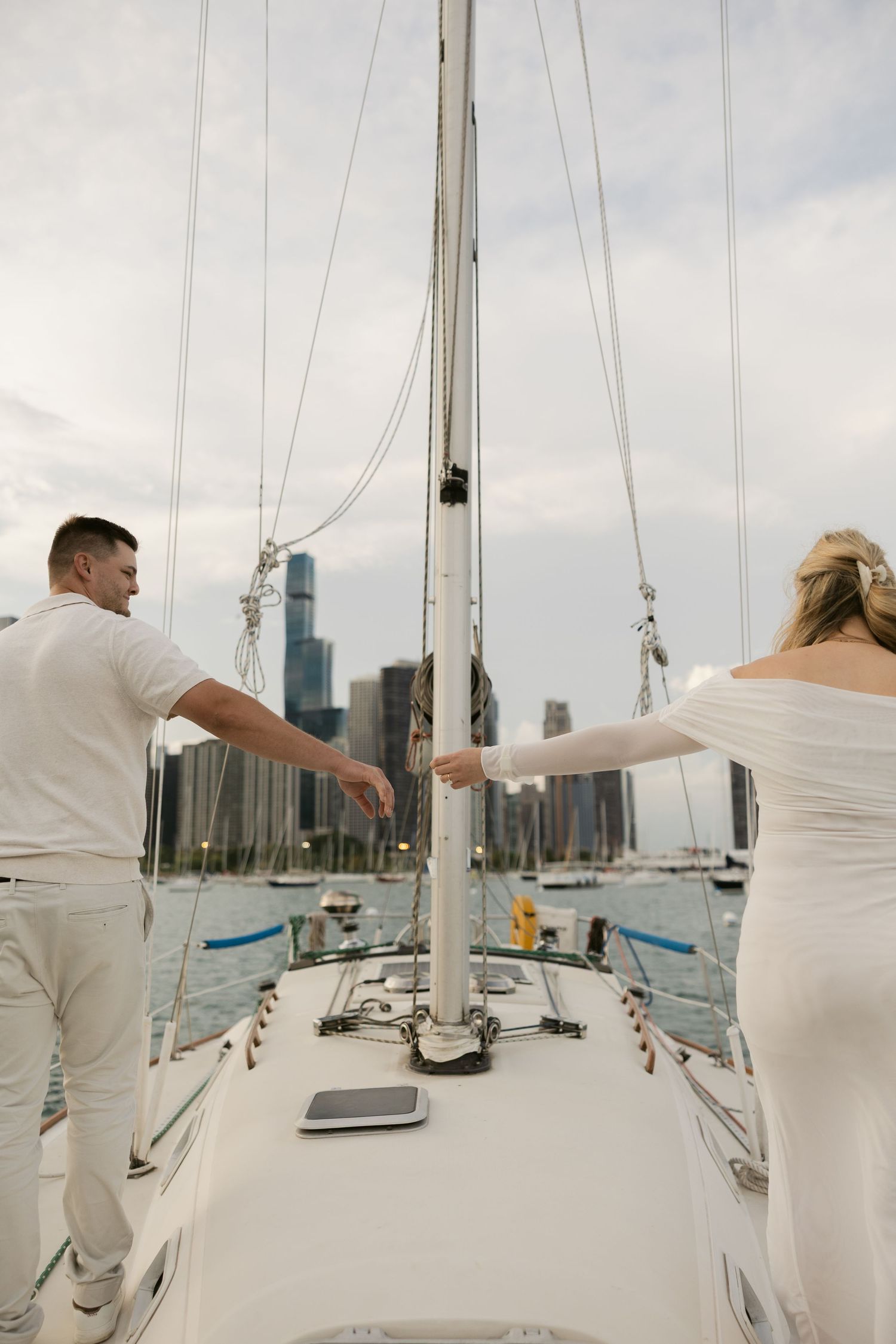 A couple in white outfits stands at the mast of their sailboat facing the city skyline.