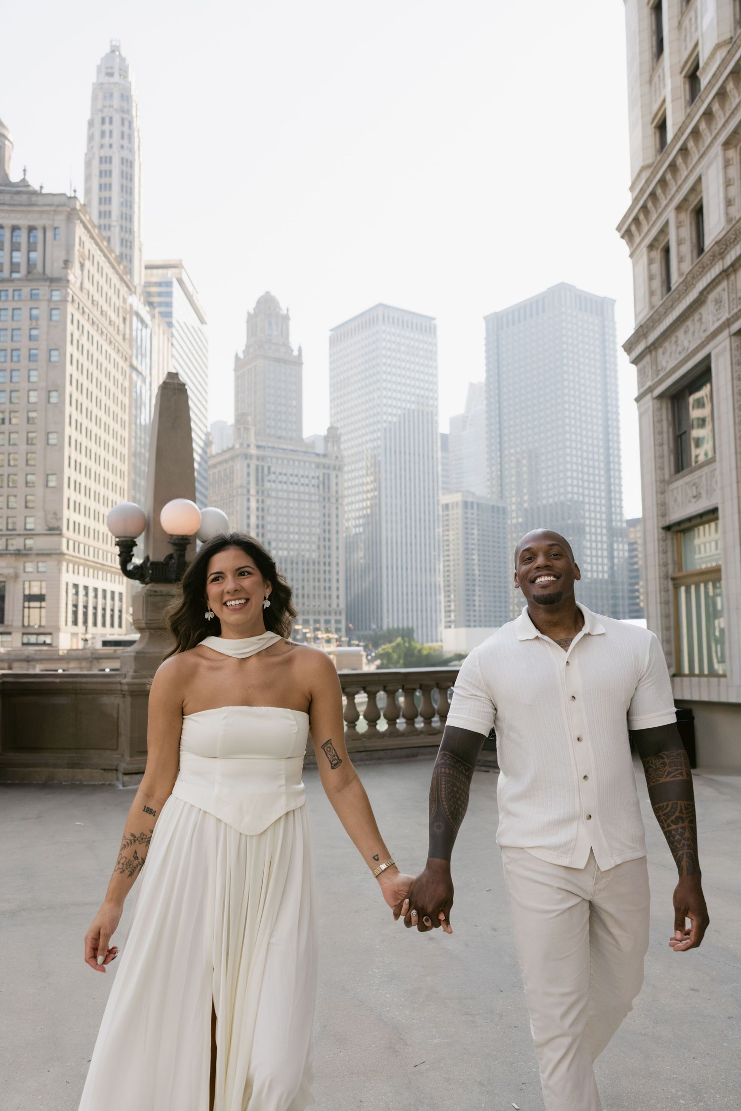 A couple in matching white outfits walks hand in hand with the Chicago skyline behind them.