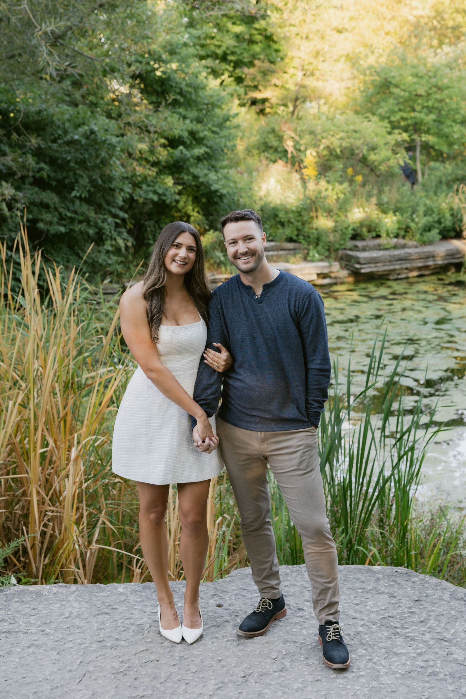A couple stands together on a stone path near a pond with water lilies and tall grasses at sunset.