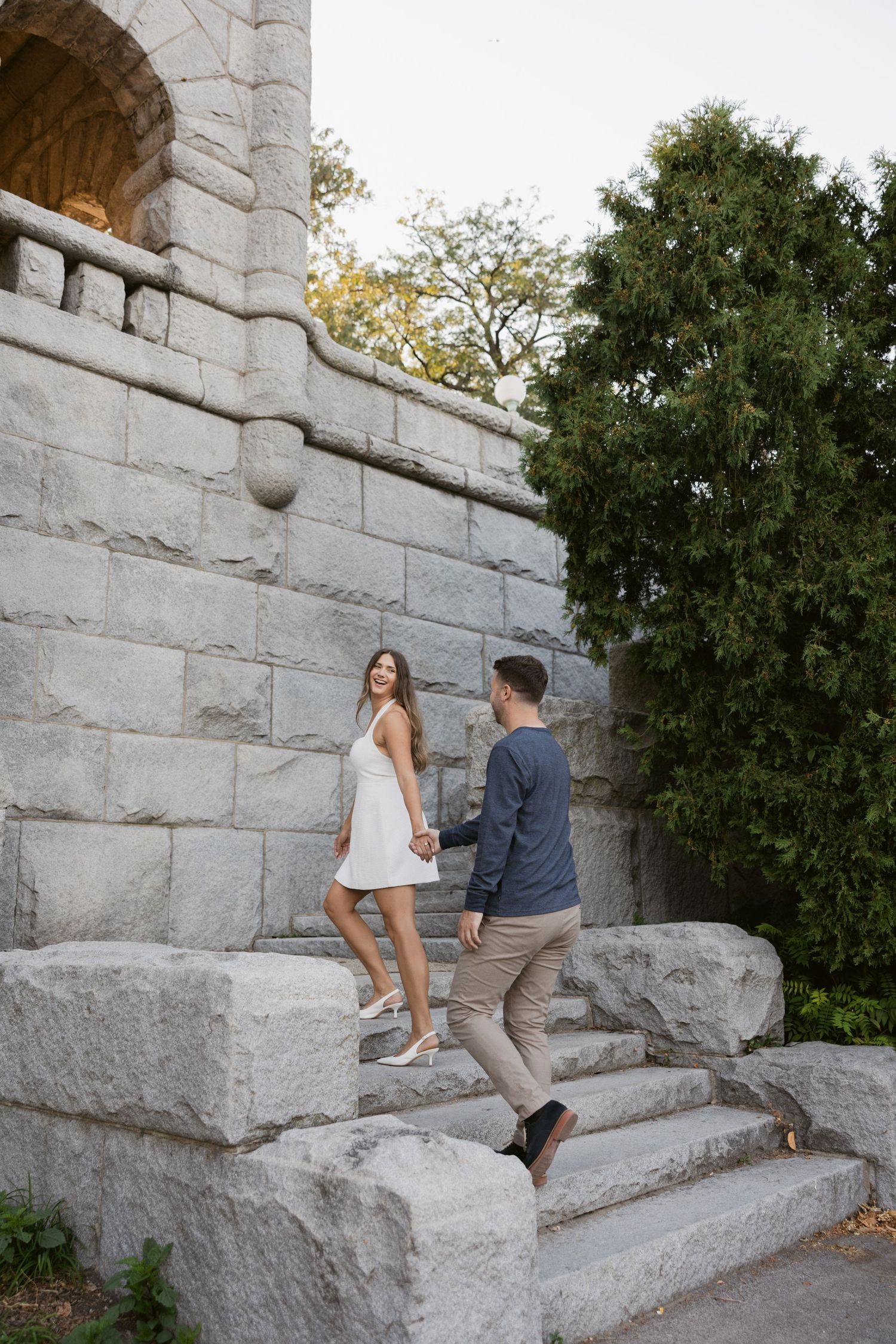 Couple playfully climbs stone steps in an outdoor garden setting.