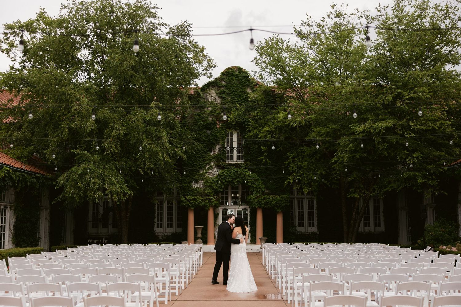 A panoramic view of an outdoor wedding ceremony setup with white chairs arranged in rows under green trees.