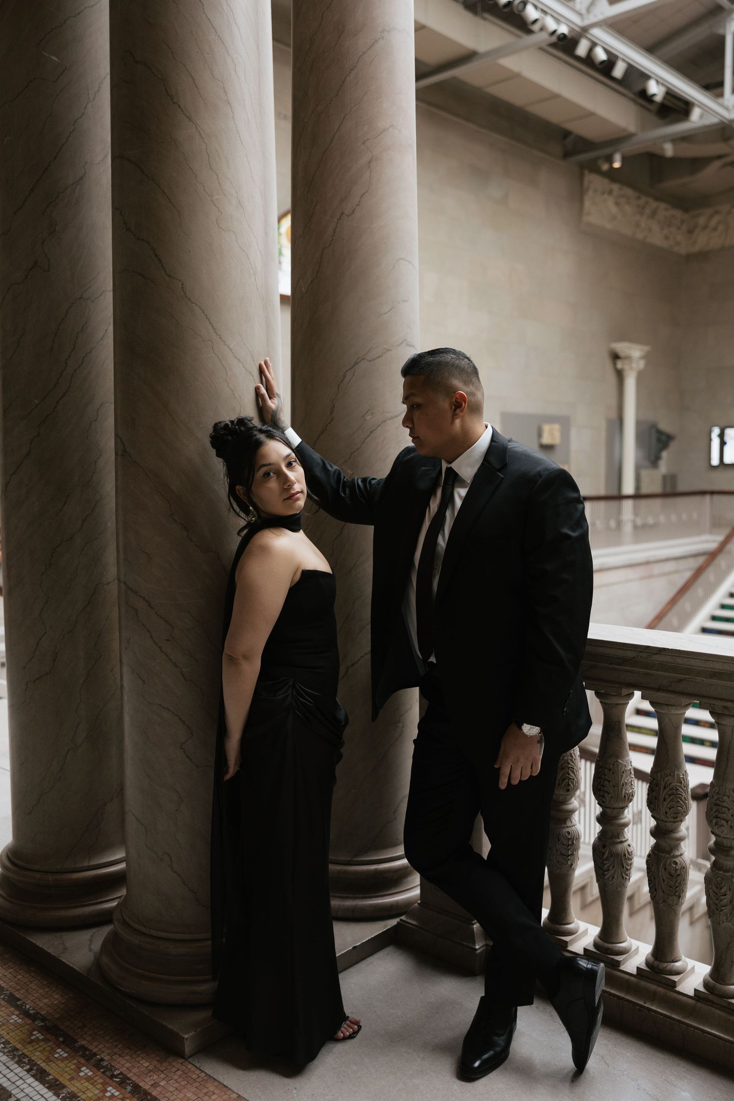 Couple in formal attire poses between classical stone columns in an elegant building.