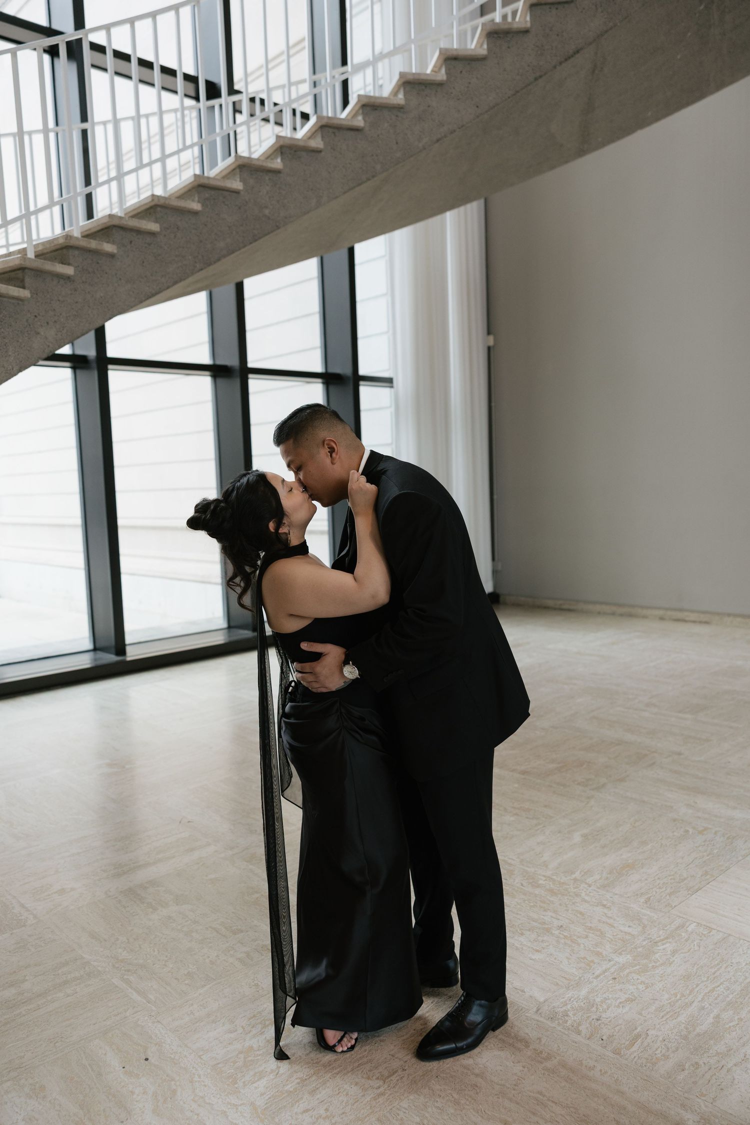 A couple in formal attire share an intimate moment against a bright window in a modern building interior.