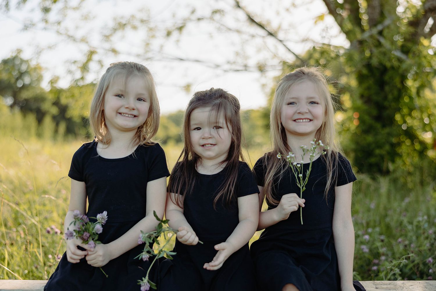 Three sisters in matching black dresses hold flowers while sitting outdoors.