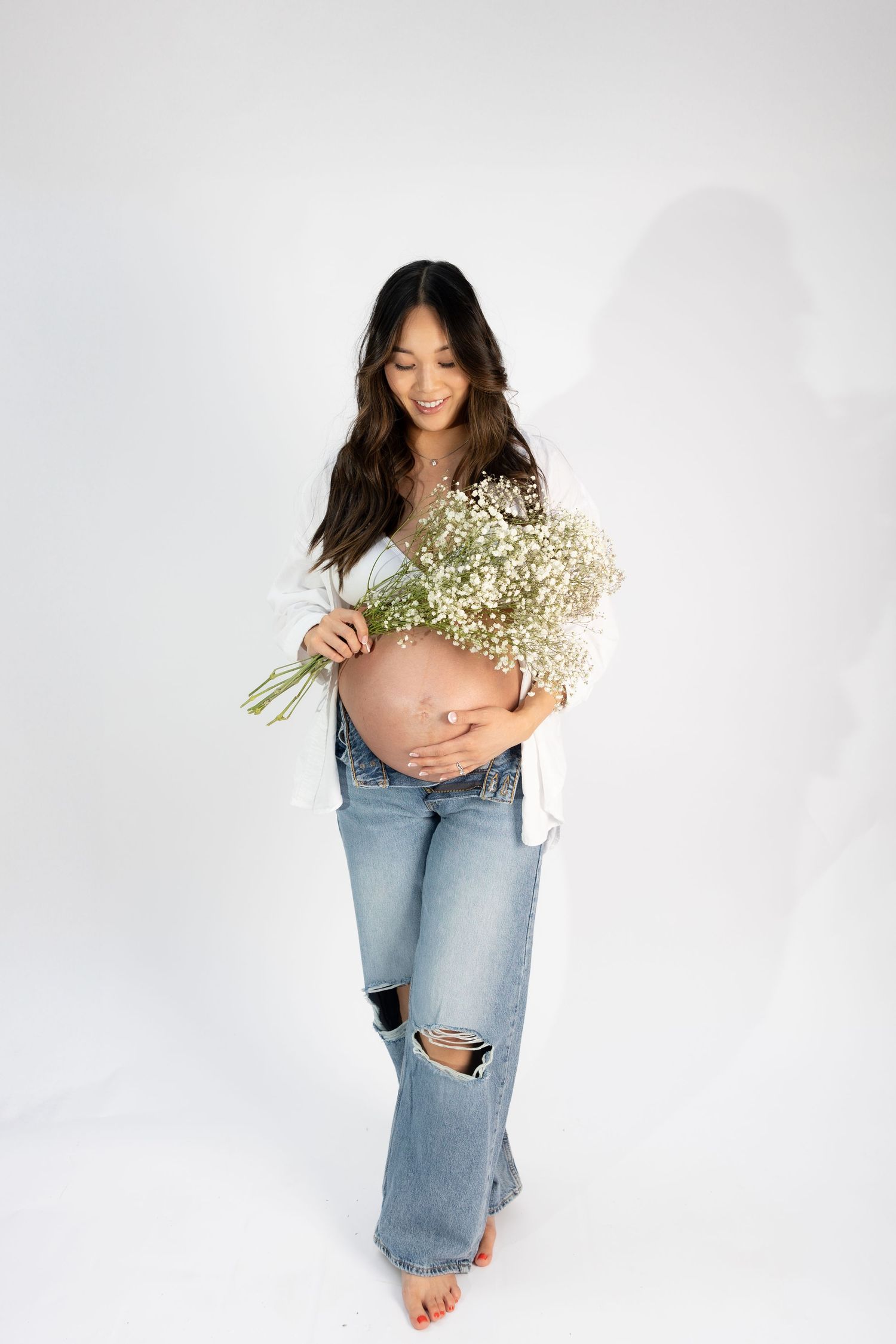 Minimalist maternity photo with woman holding baby's breath bouquet against white wall.
