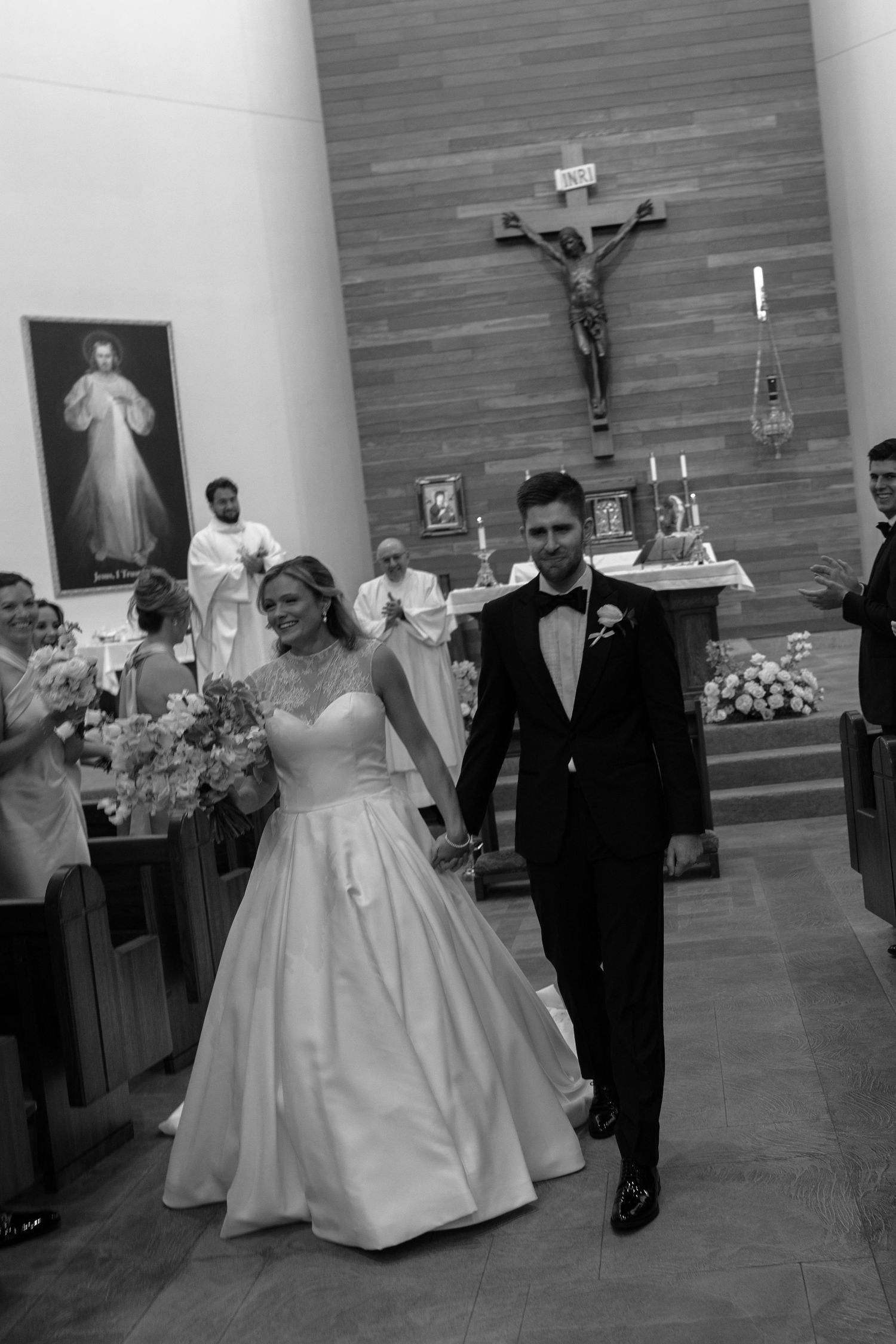 A wedding ceremony exits a modern church with crucifix and Divine Mercy painting visible in background.