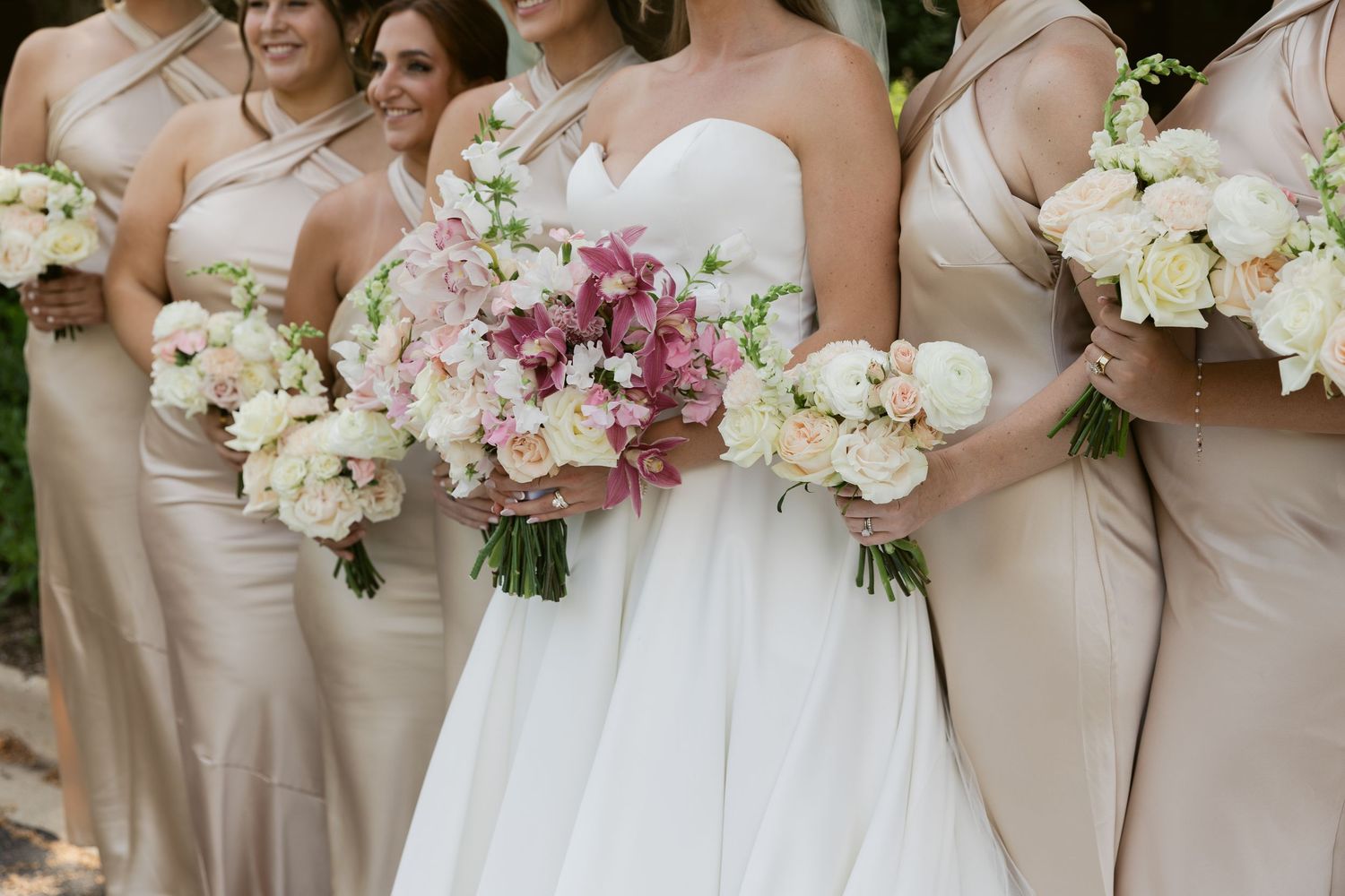 Bridesmaids in champagne dresses holding white and pink bouquets outdoors.