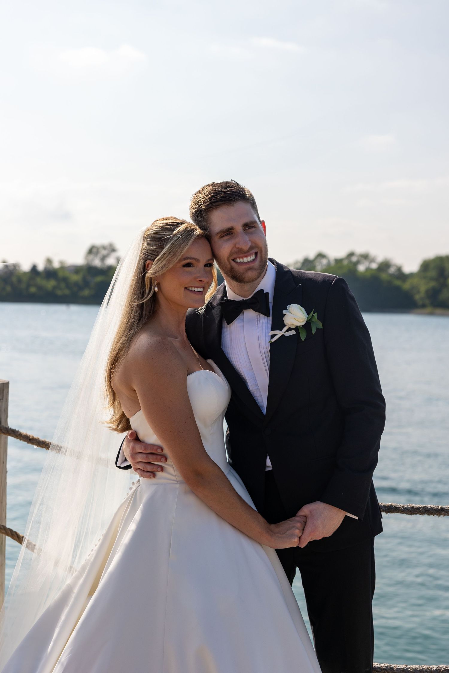A romantic wedding couple embraces and laughs together by a waterfront setting.