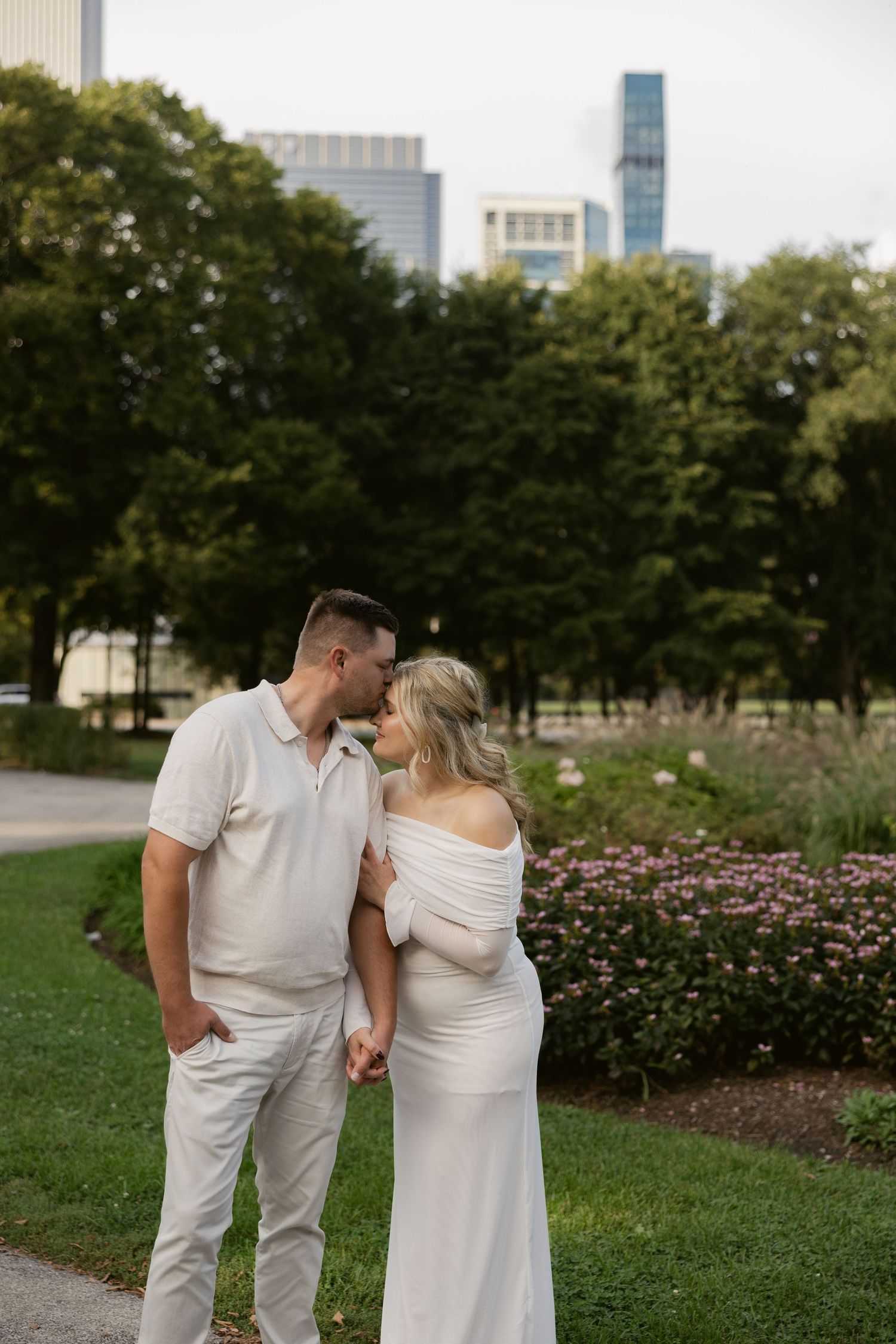 A romantic couple in all-white attire embraces in a garden setting with city buildings visible in the background.