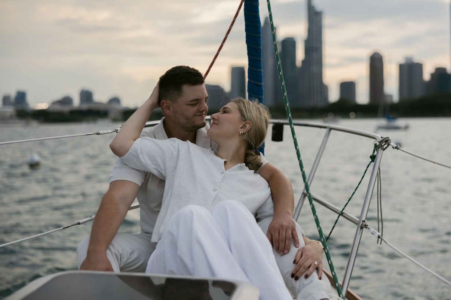 Two people dressed in white relaxing on the bow of a sailboat during sunset with skyline views.