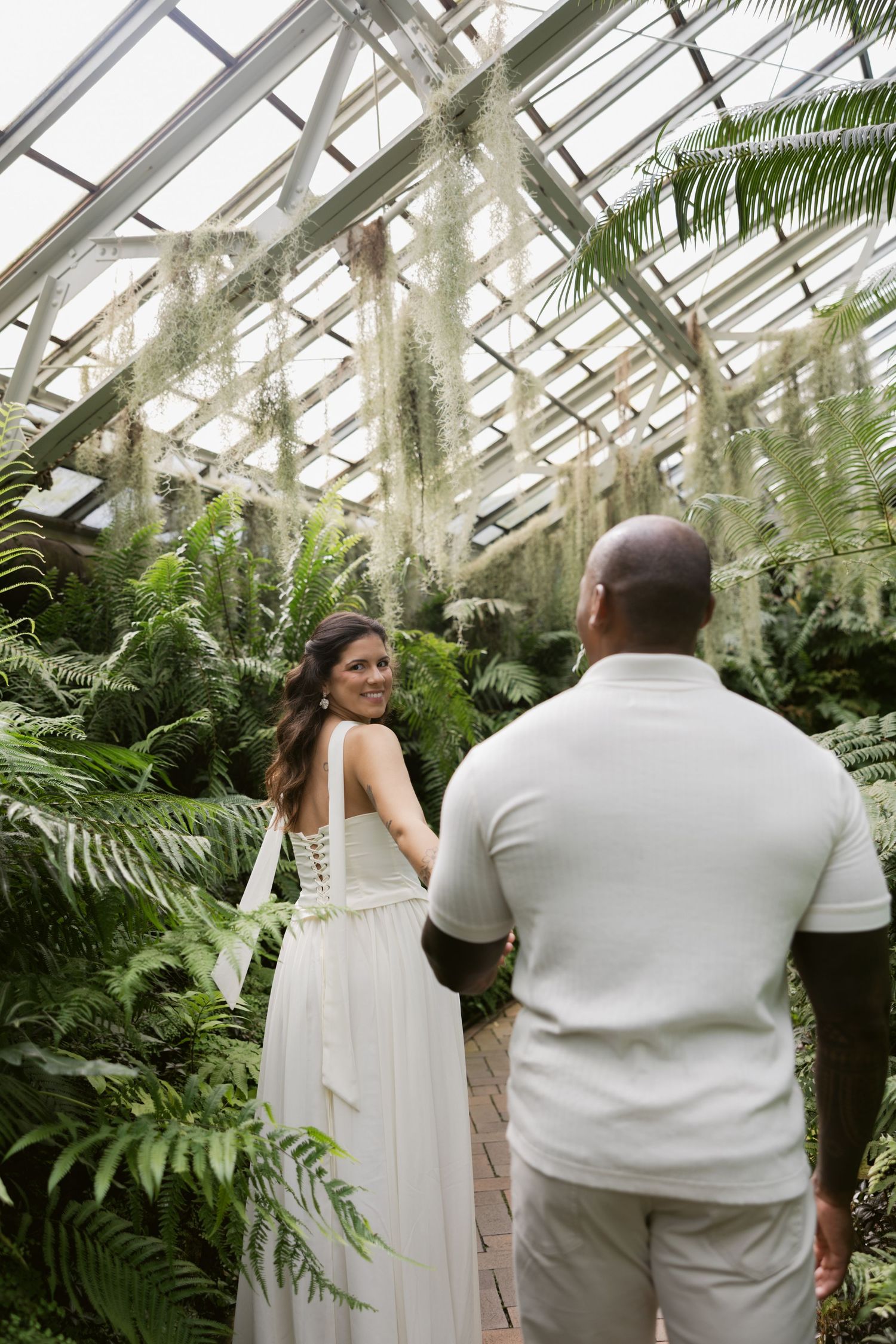 Couple in elegant white attire poses in a greenhouse filled with lush ferns and hanging plants.