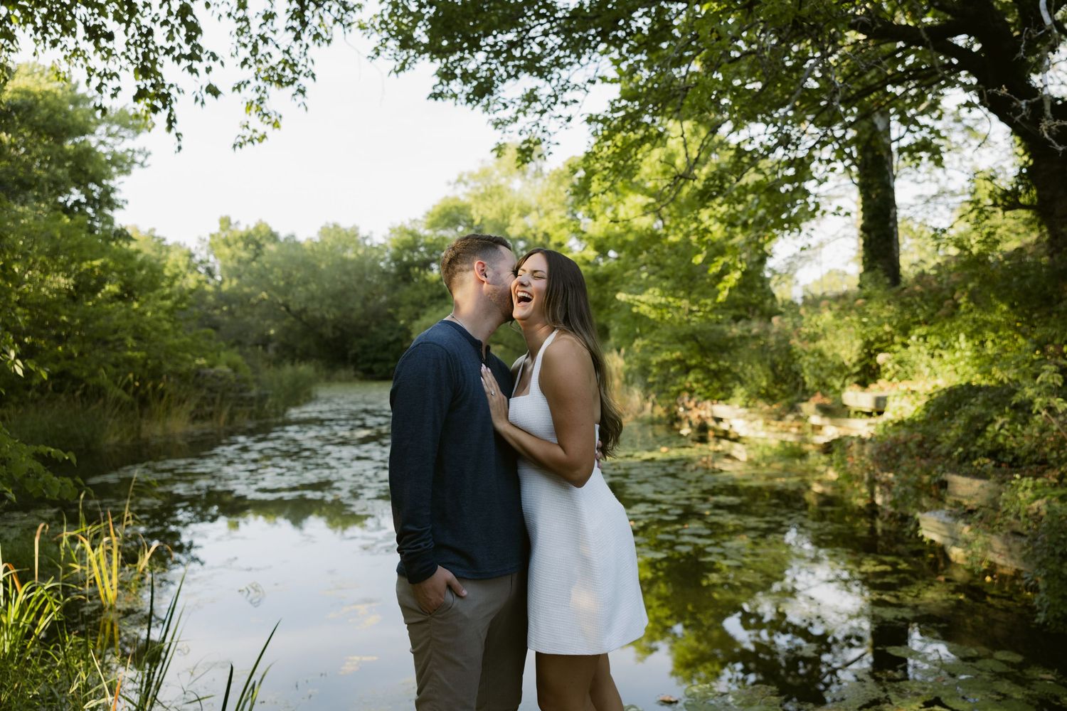 Romantic couple embraces during sunset photo session near scenic pond in wooded garden setting.