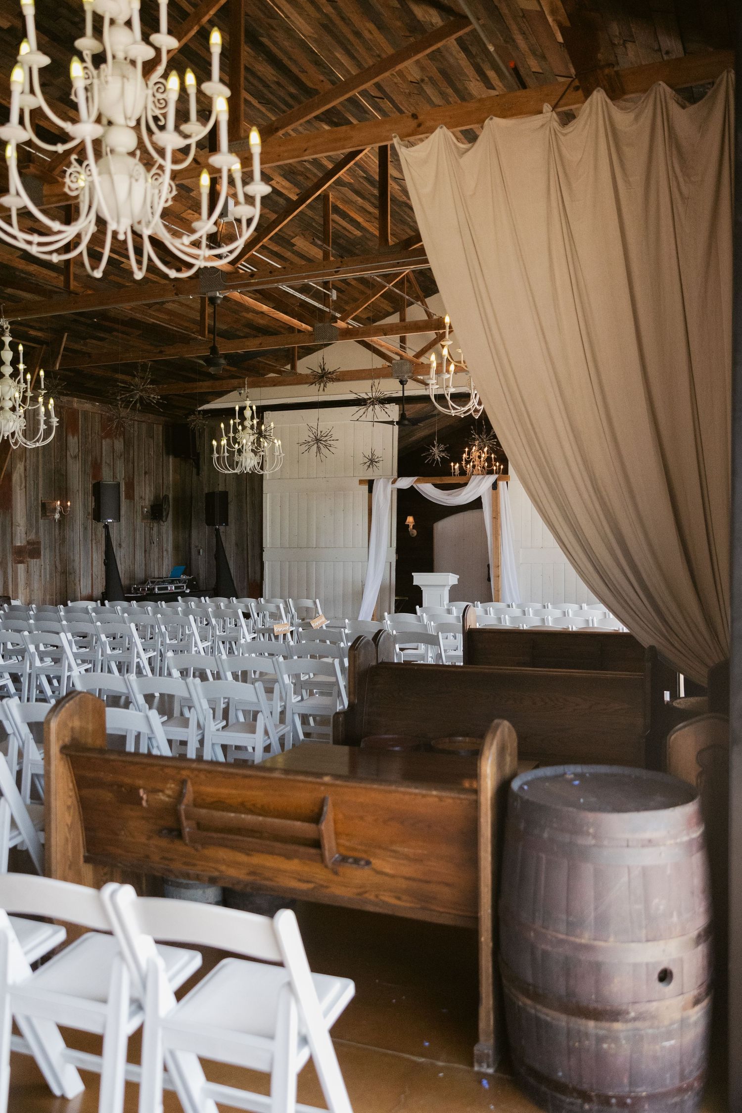 Rustic barn wedding venue with wooden benches, chandeliers, hanging drapes, and decorative wine barrels.