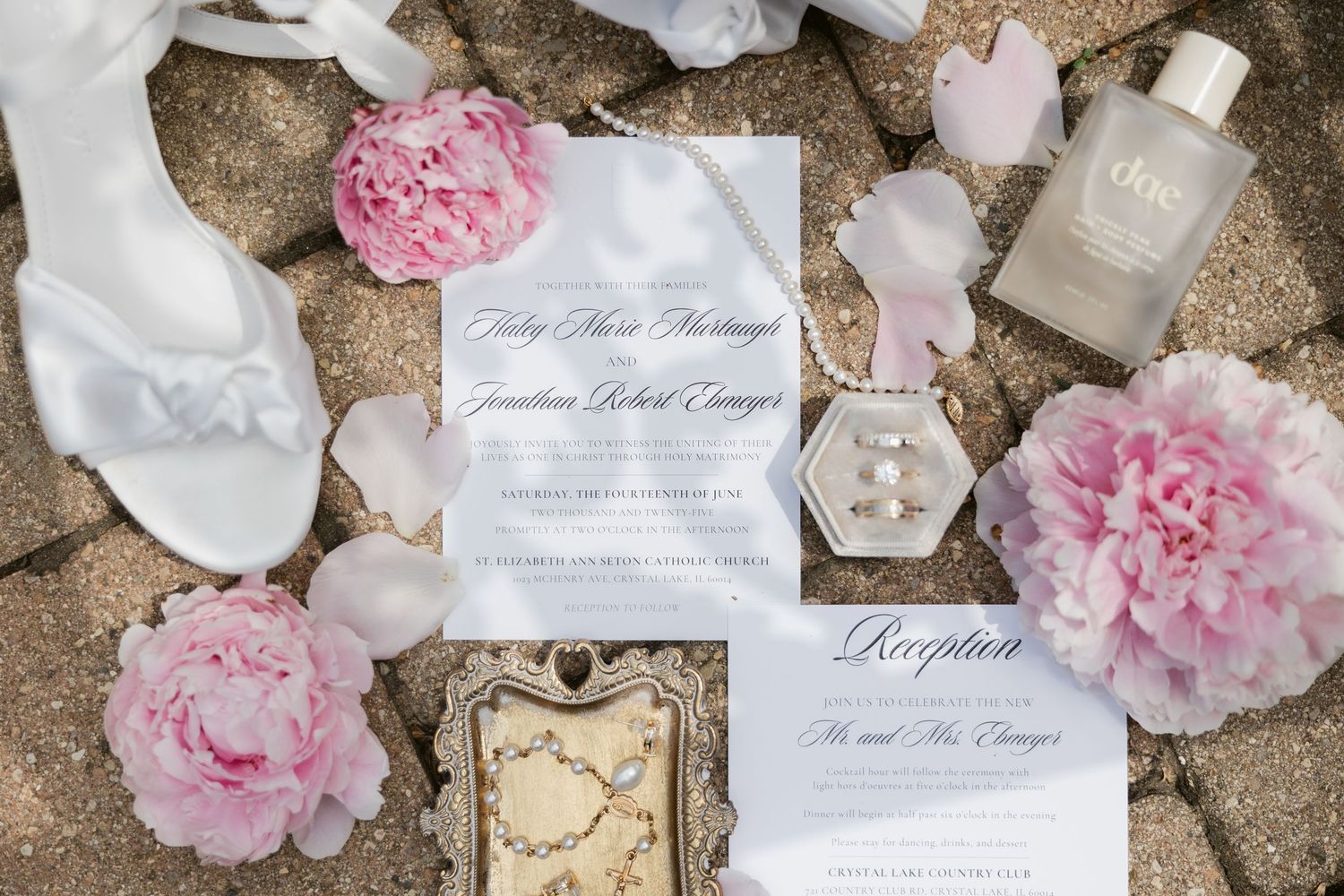 Wedding details flatlay with pink peonies, white wedding invitation, perfume bottle and bridal shoes on stone surface.