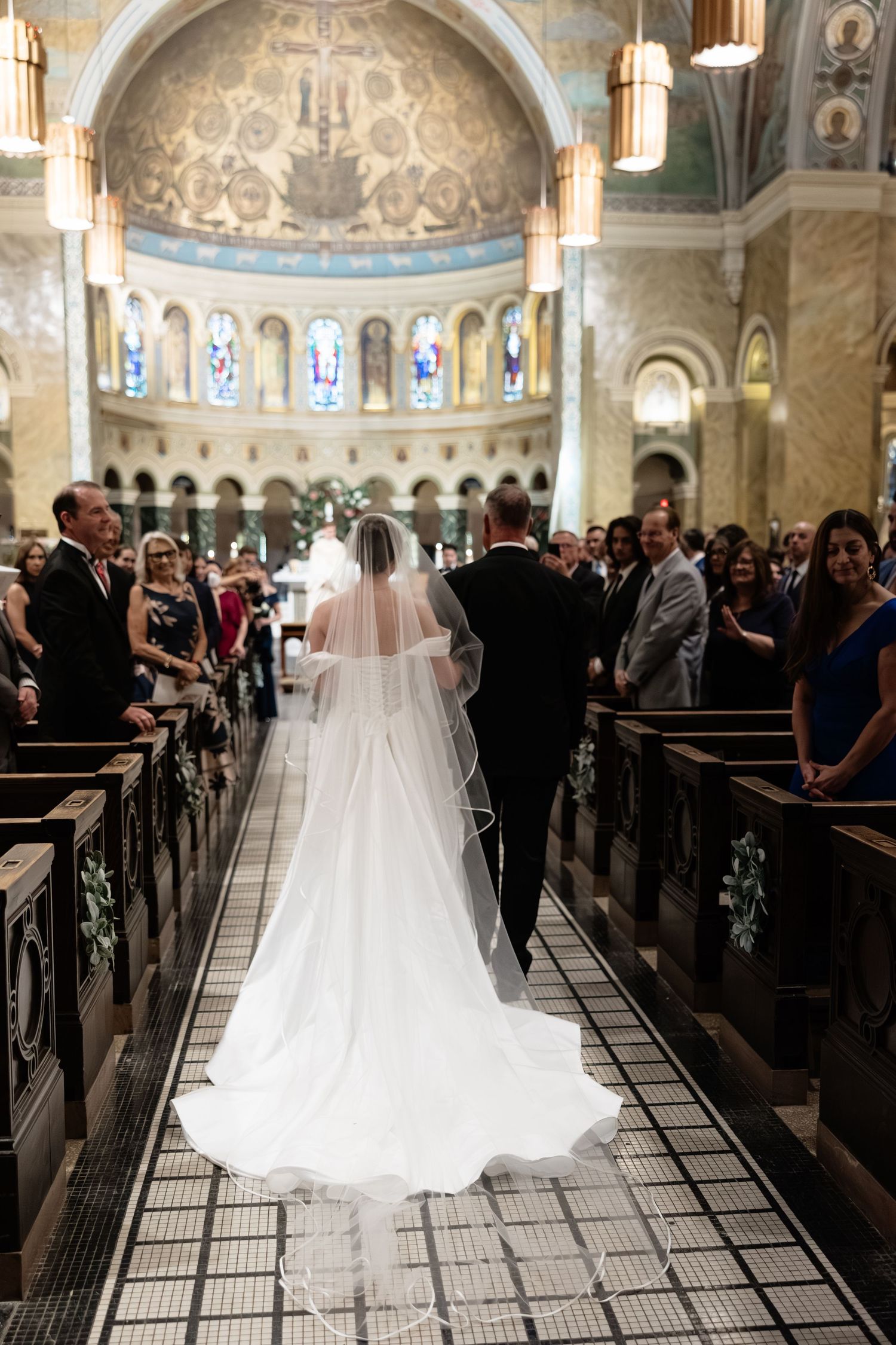 A bride walks down the aisle in a cathedral with ornate domed ceilings and stained glass windows during a wedding ceremony.