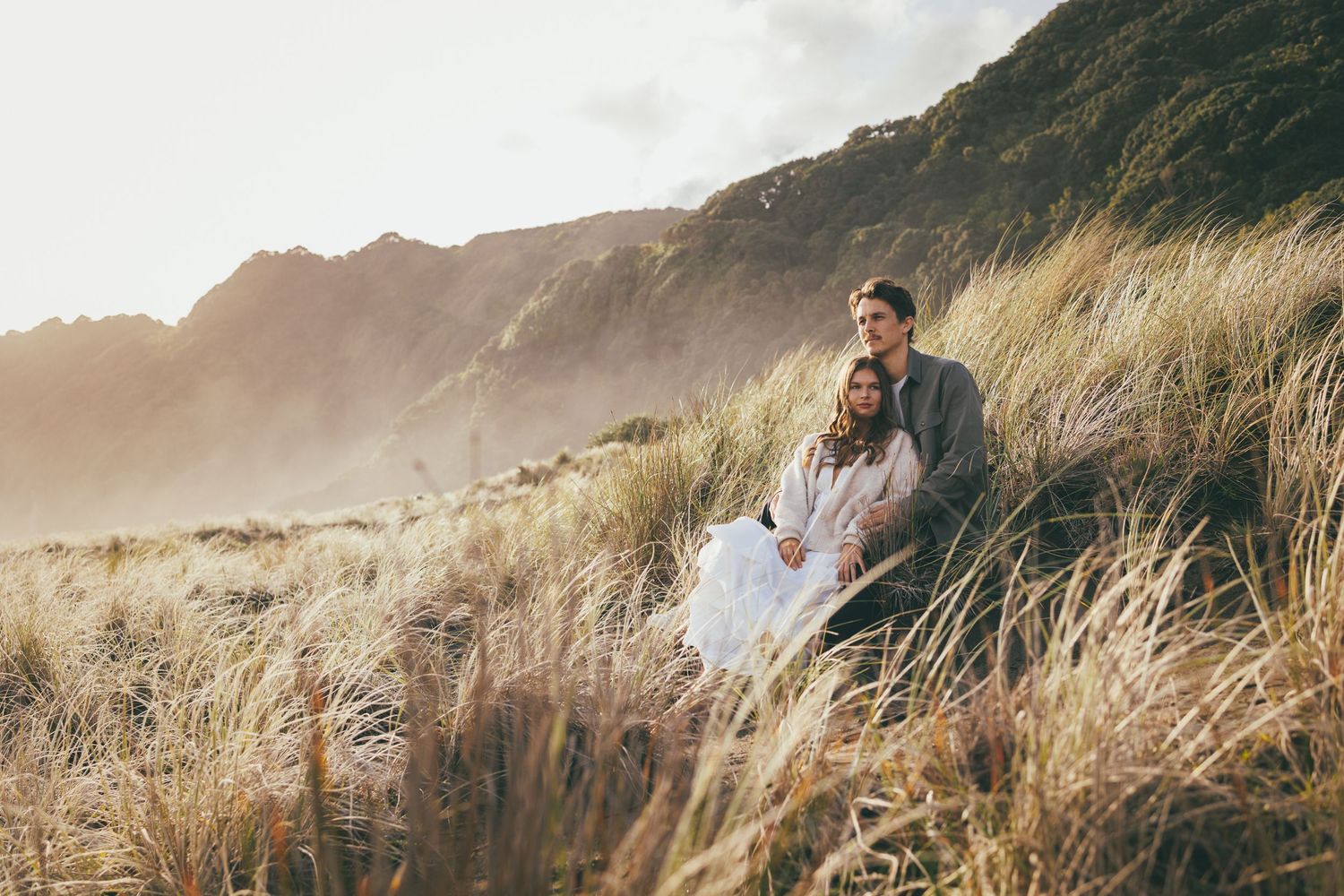 Couple sitting together in golden coastal dunes with misty mountains, romantic outdoor portrait in natural light.