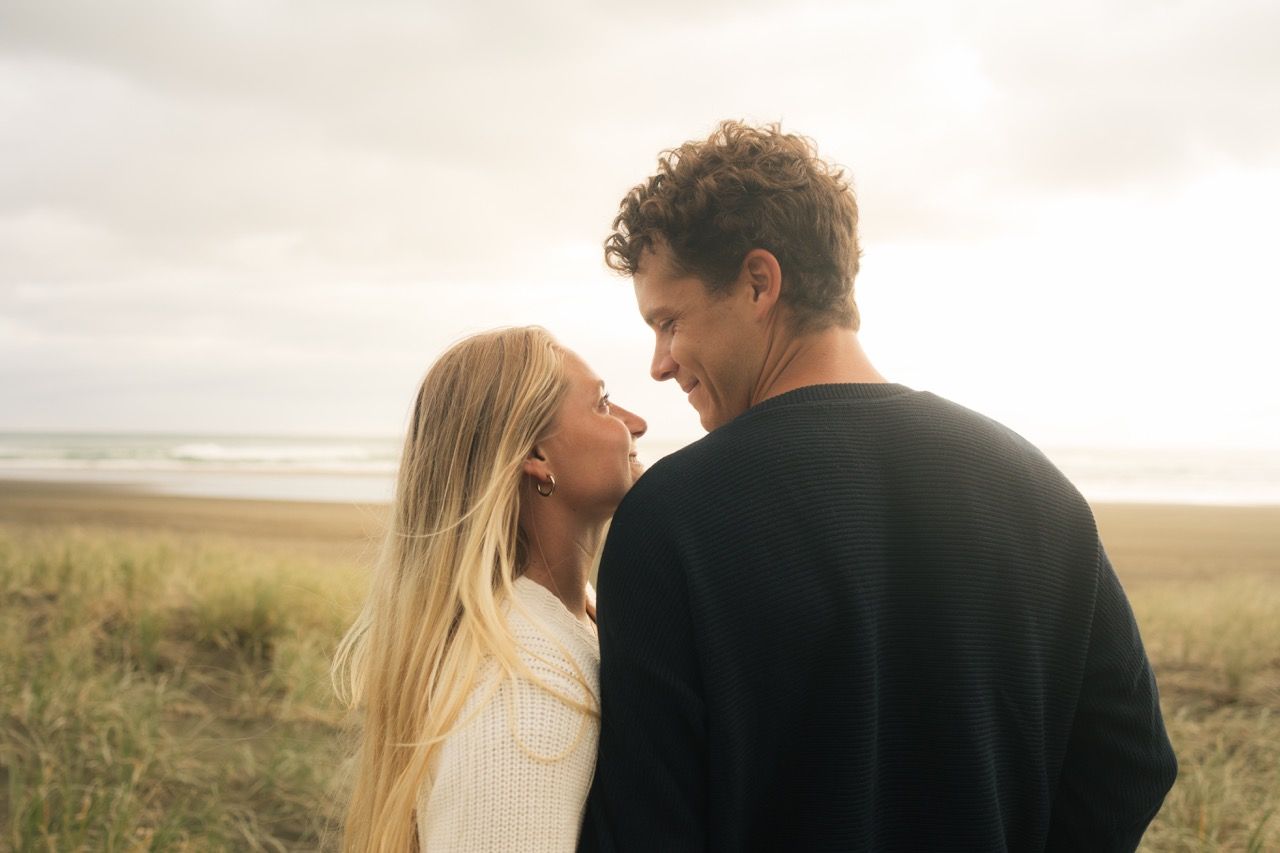 Romantic couple sharing a tender moment on a beach, with ocean waves and golden grass in the background.