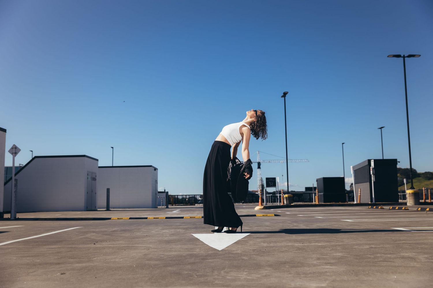 Woman in white top and black wide-leg pants leaning forward in sunny parking lot holding black bag.