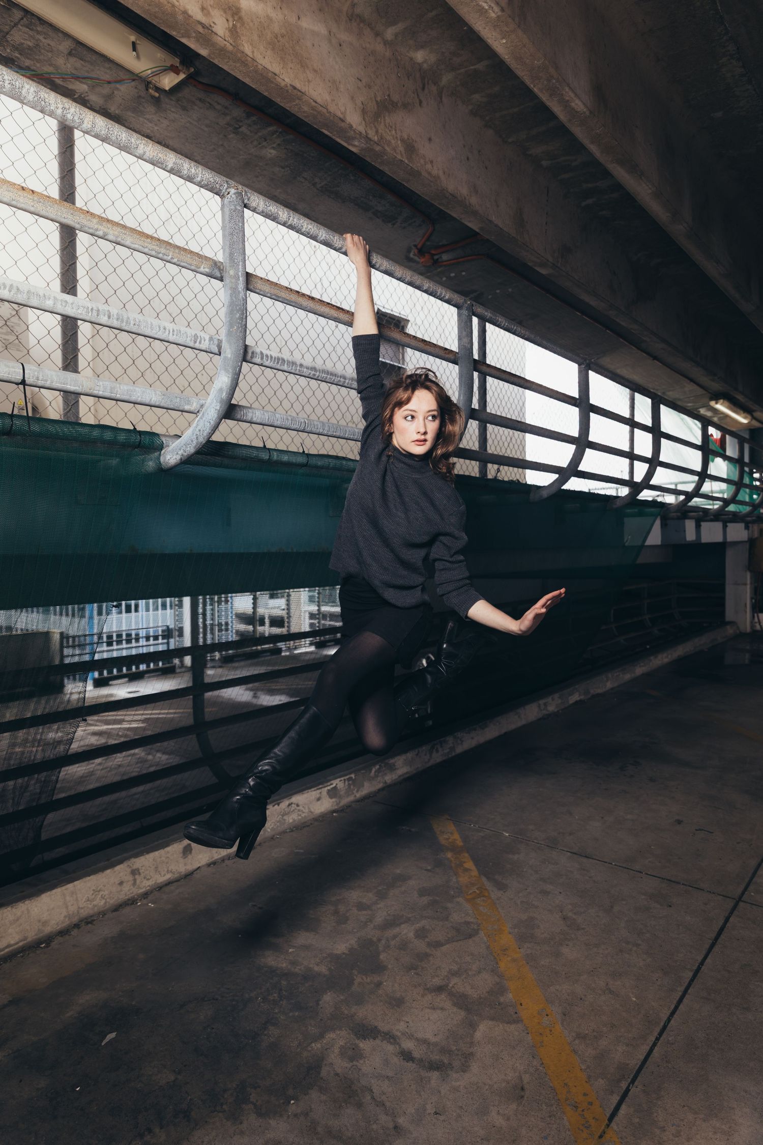 Young woman in black outfit hanging from fence in urban parking structure, one leg extended sideways.
