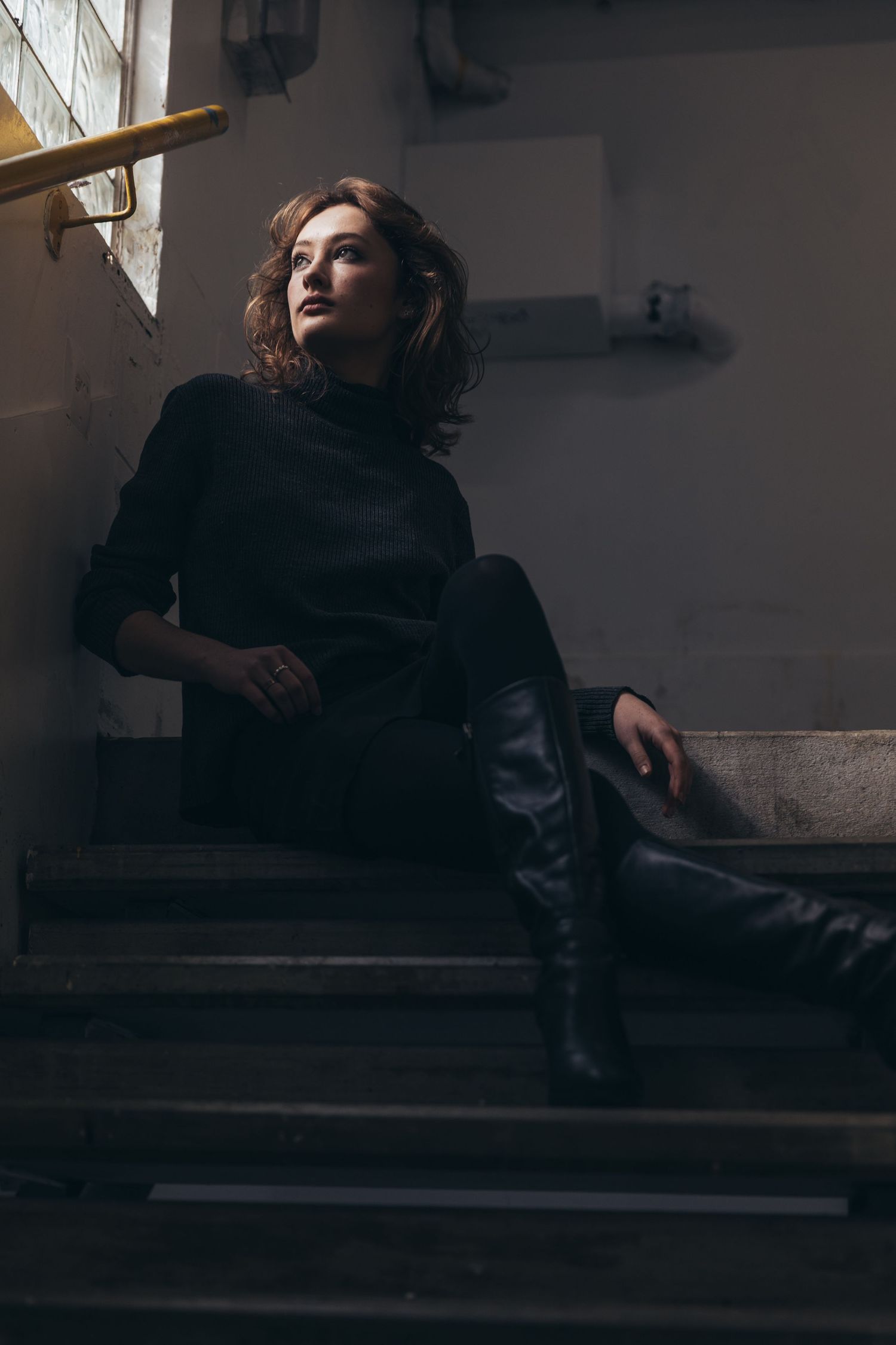 Woman in black turtleneck and knee-high boots sitting on dimly lit concrete staircase.
