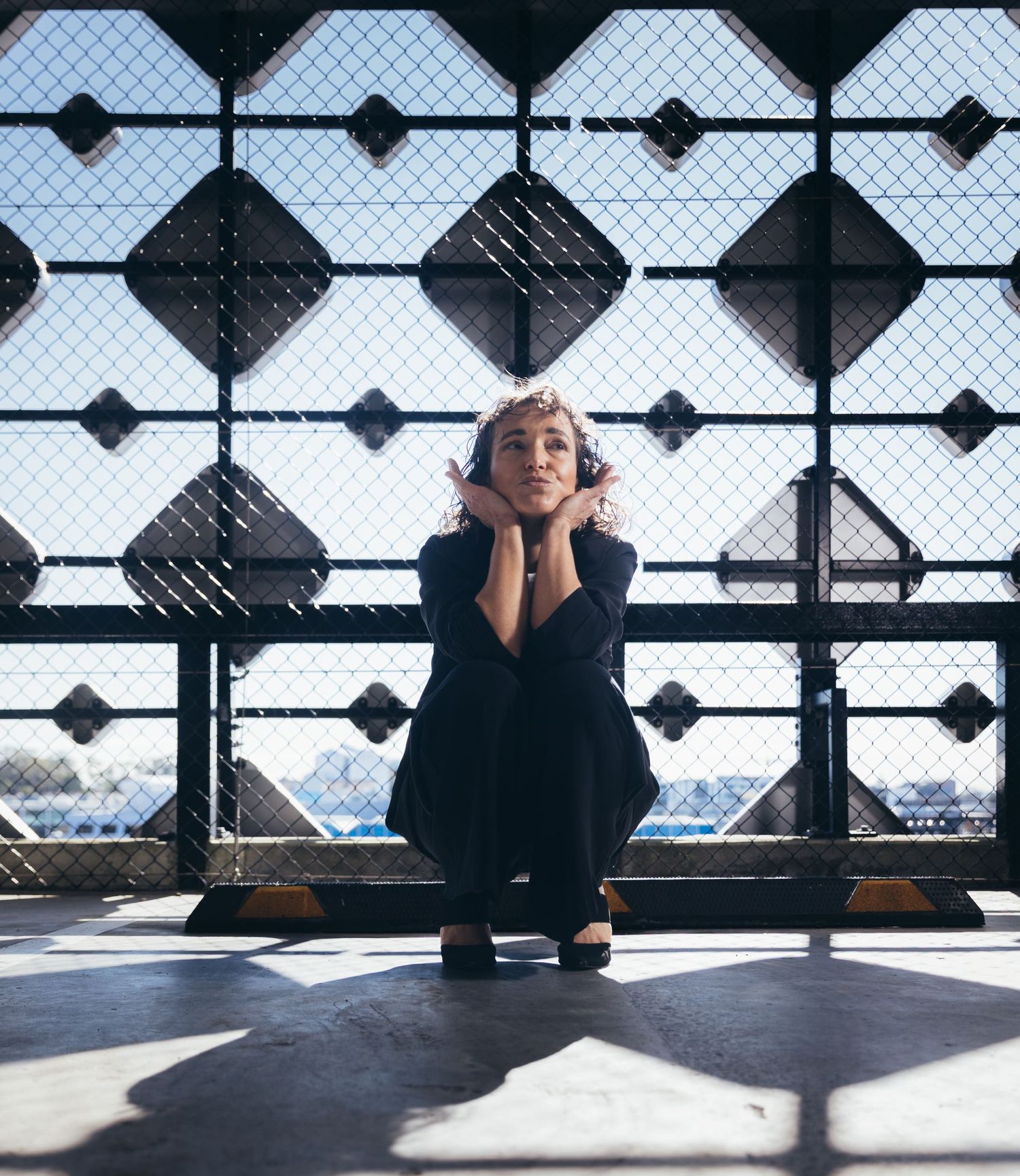 Woman in all-black outfit crouching with hands under chin against geometric metal fence in sunlight.