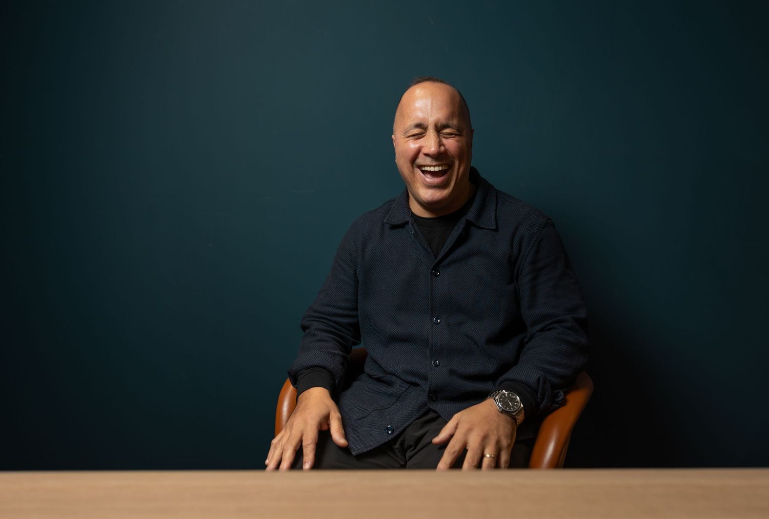 Laughing man in a navy shirt seated in a leather chair against a dark teal background.