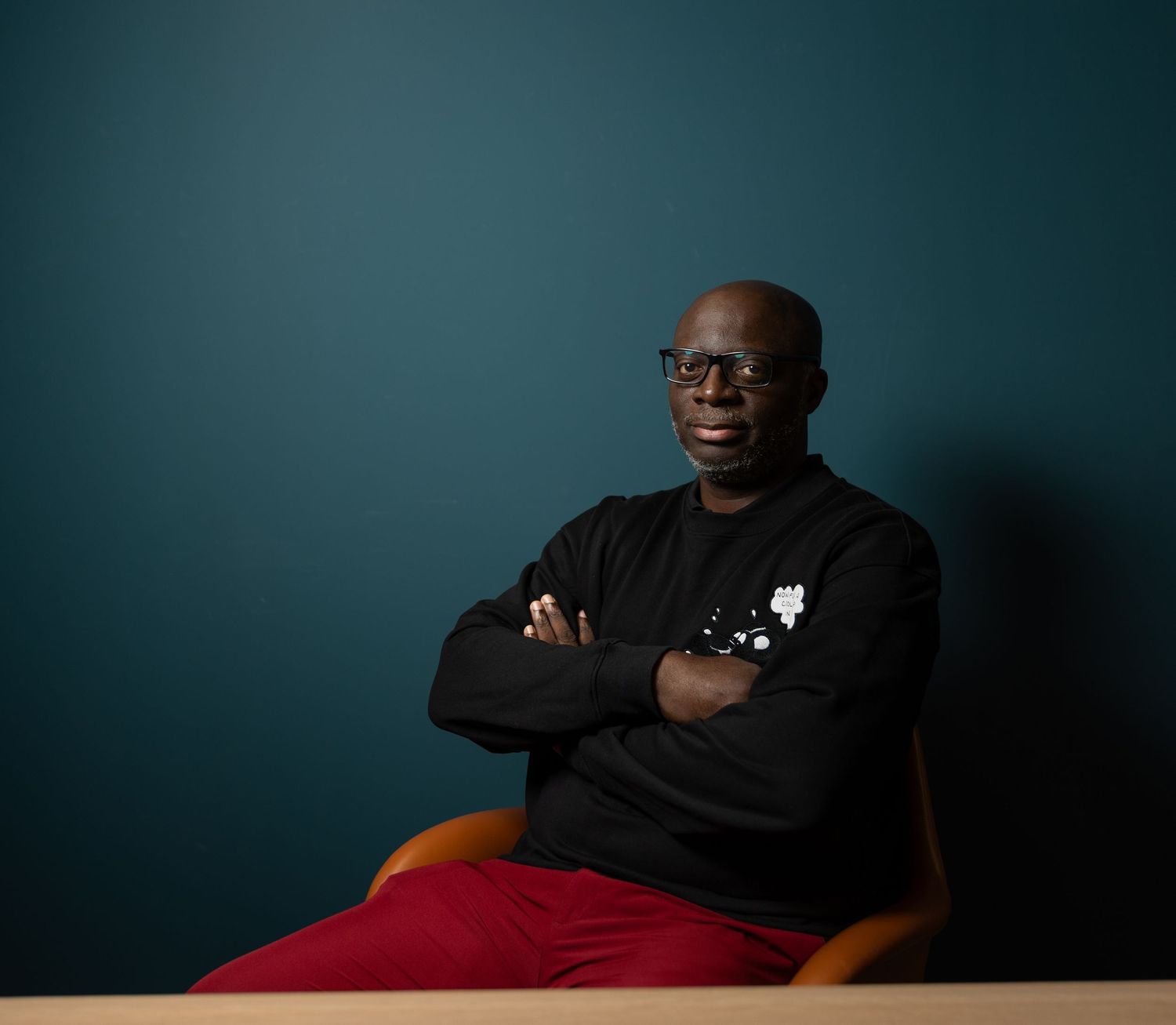 Confident man with glasses in a black sweatshirt and red pants sitting against a teal wall.