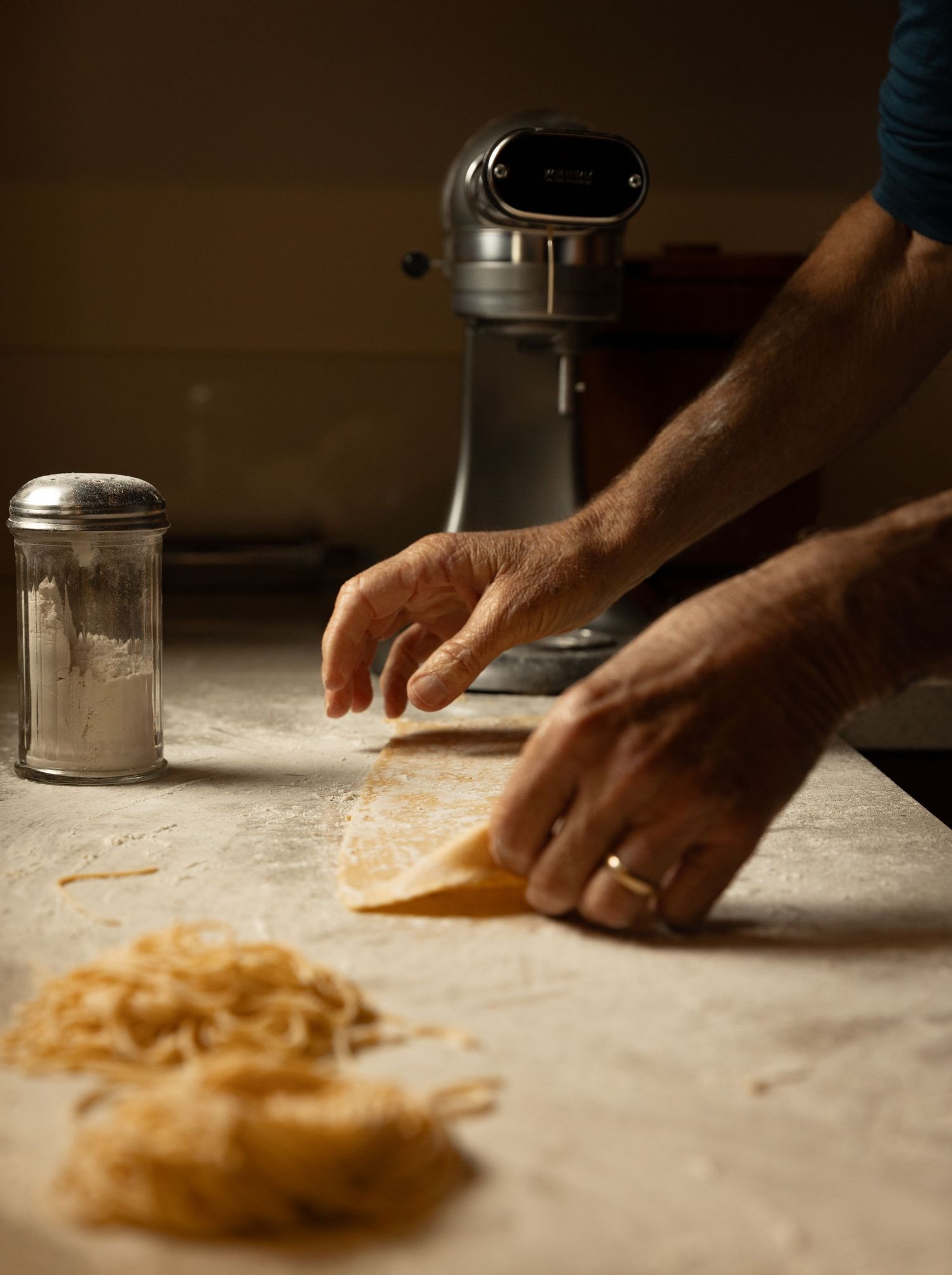 Hands rolling fresh pasta dough on a floured counter beside a KitchenAid mixer.