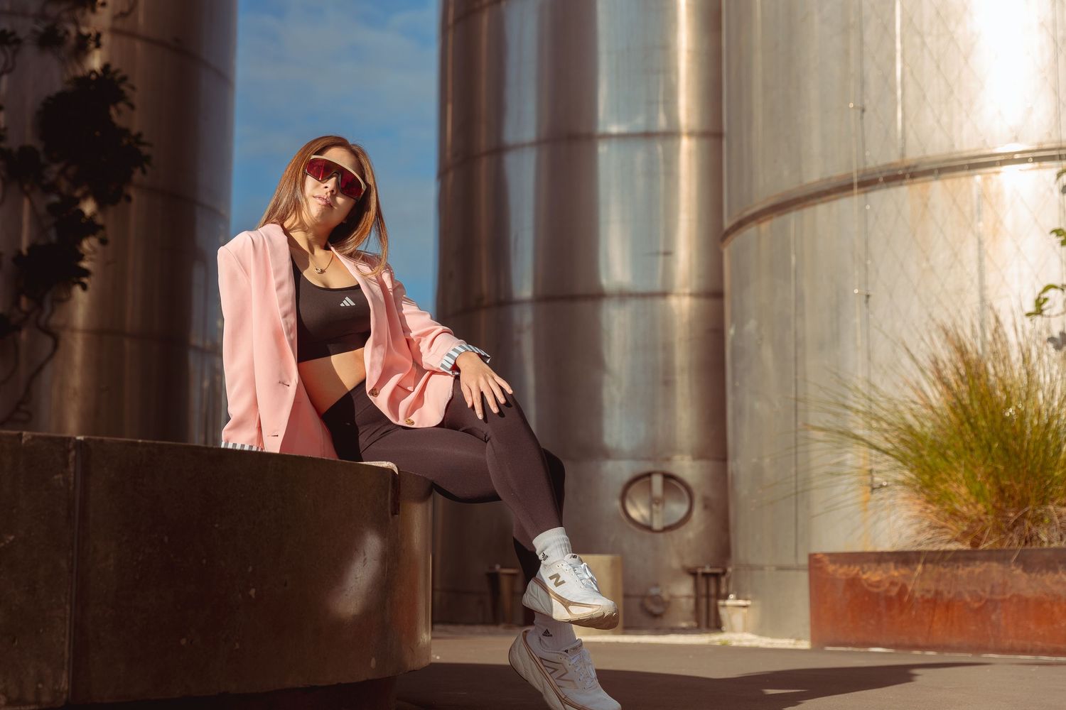 Woman in pink blazer and black activewear sitting relaxed outdoors near industrial silos in sunlight.