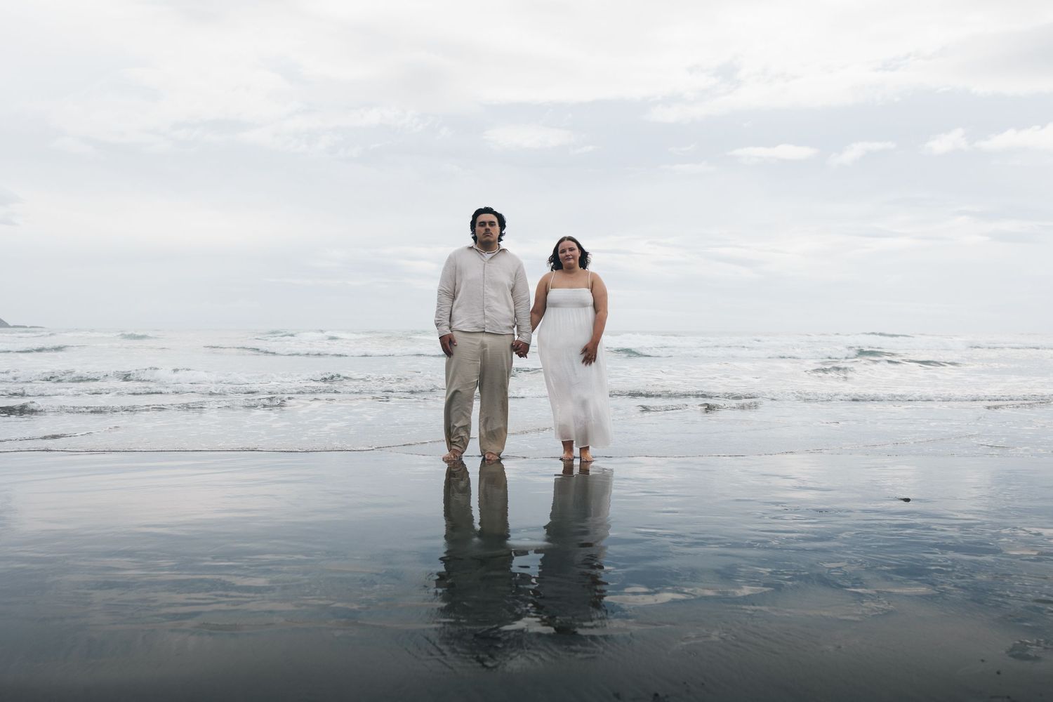 Couple holding hands standing in shallow ocean water on a cloudy beach day.