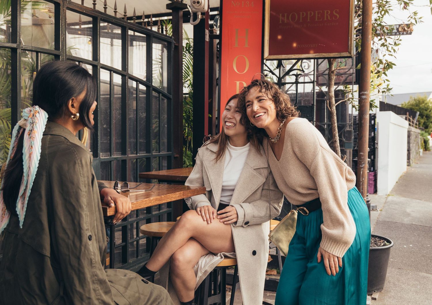 Three women laughing and socializing outside a restaurant cafe on a city street.