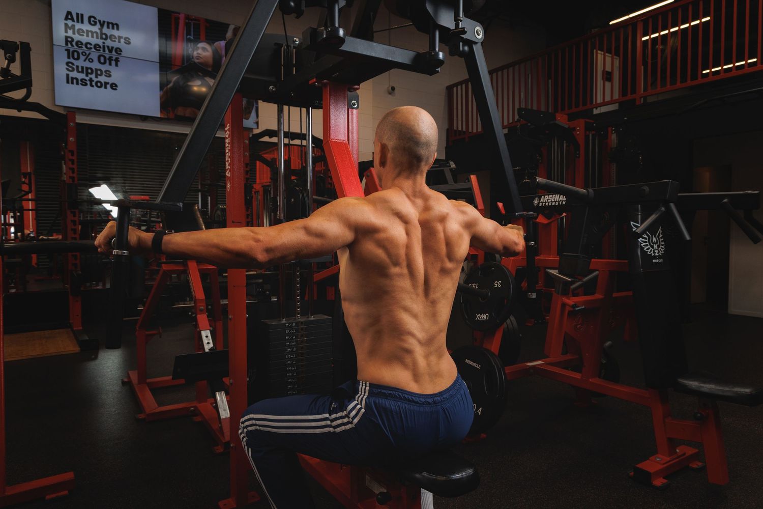 Muscular bald man performing cable fly exercise on gym machine, showing defined back muscles in blue shorts.