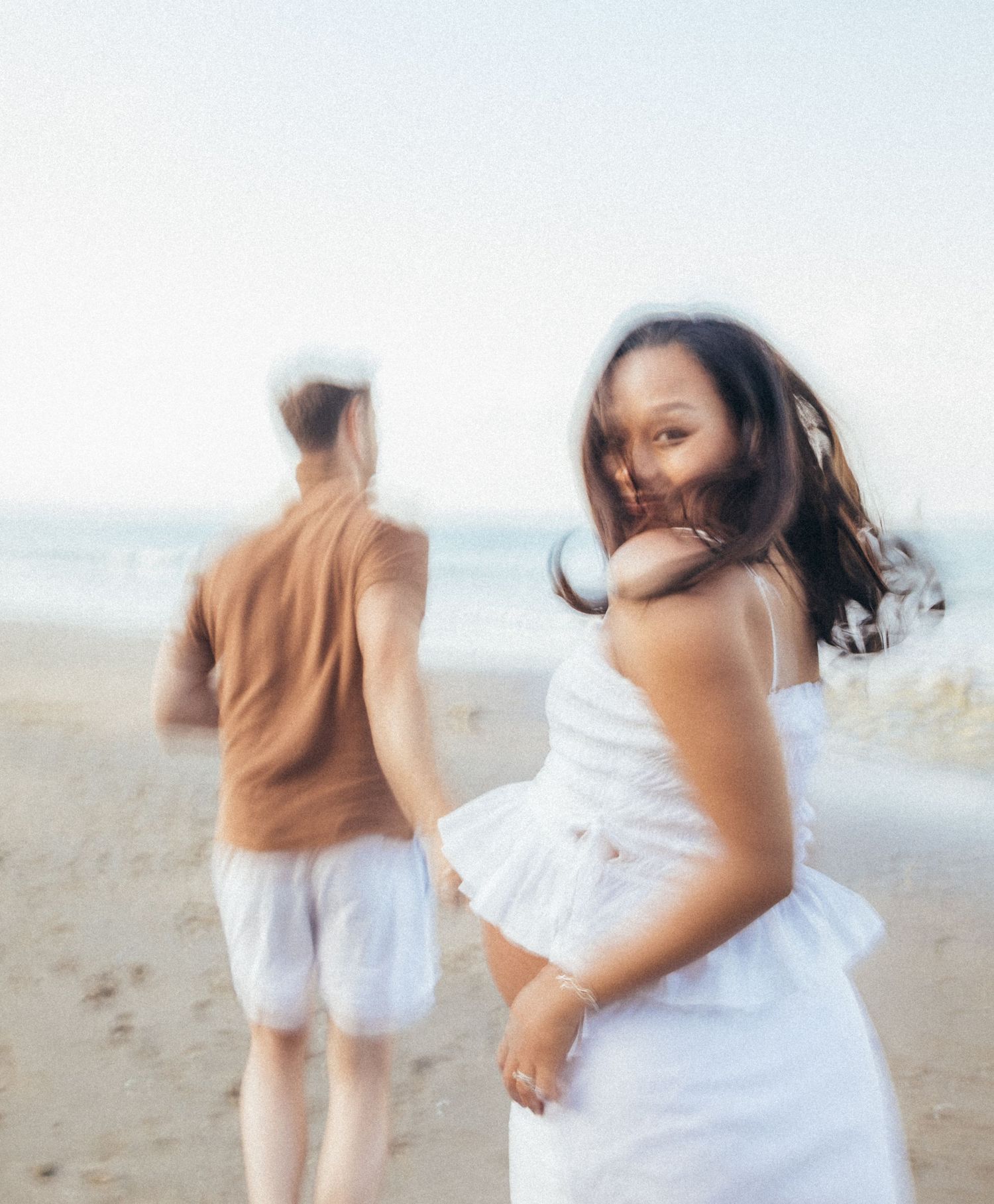A woman in a white dress smiles on a beach while a man in brown shirt and white shorts walks ahead.