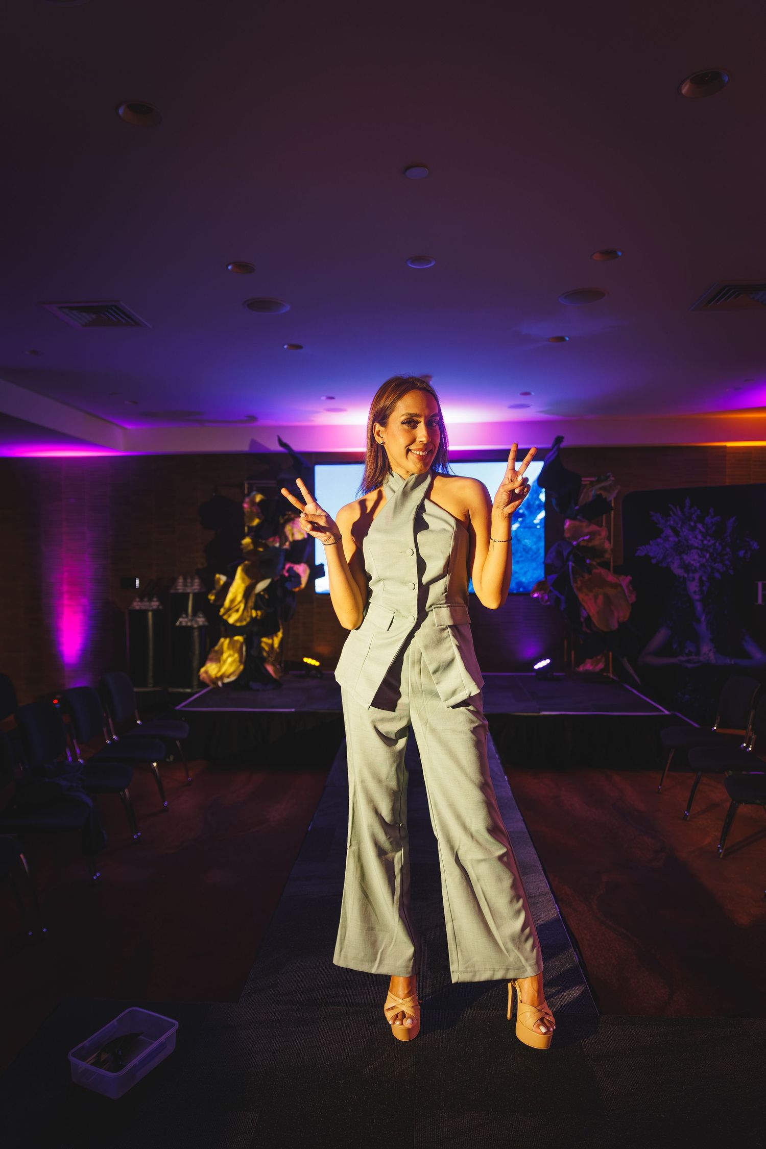 Confident woman in grey halter jumpsuit posing with peace signs on an event stage with purple lighting.