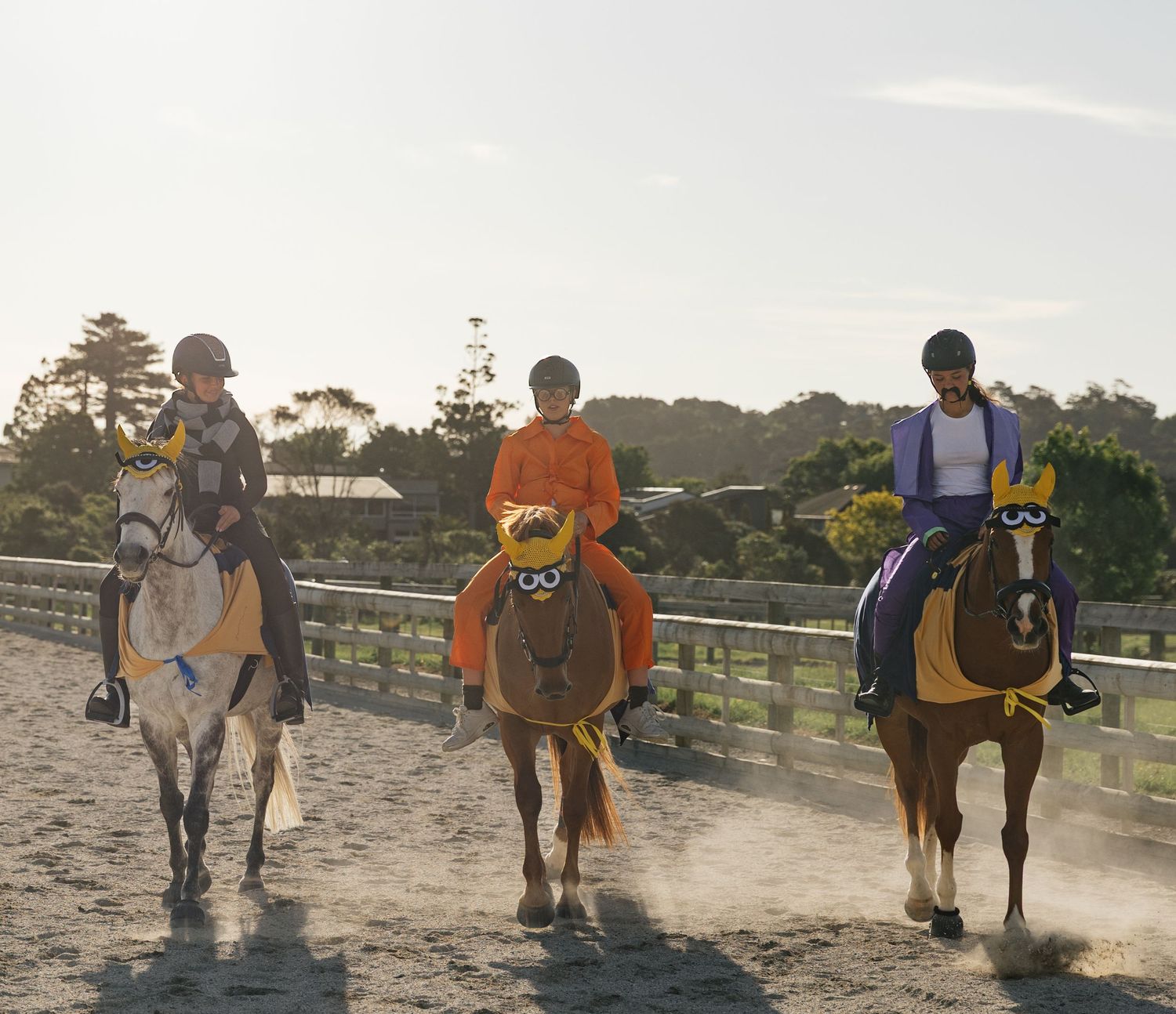 Three riders on horses walking through dusty equestrian arena in golden evening sunlight.
