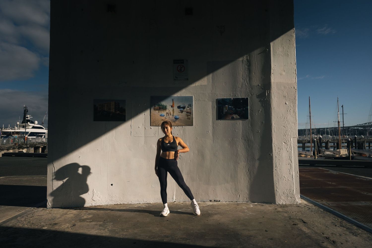 Athletic woman in black activewear posing beside a concrete wall at a sunny marina.