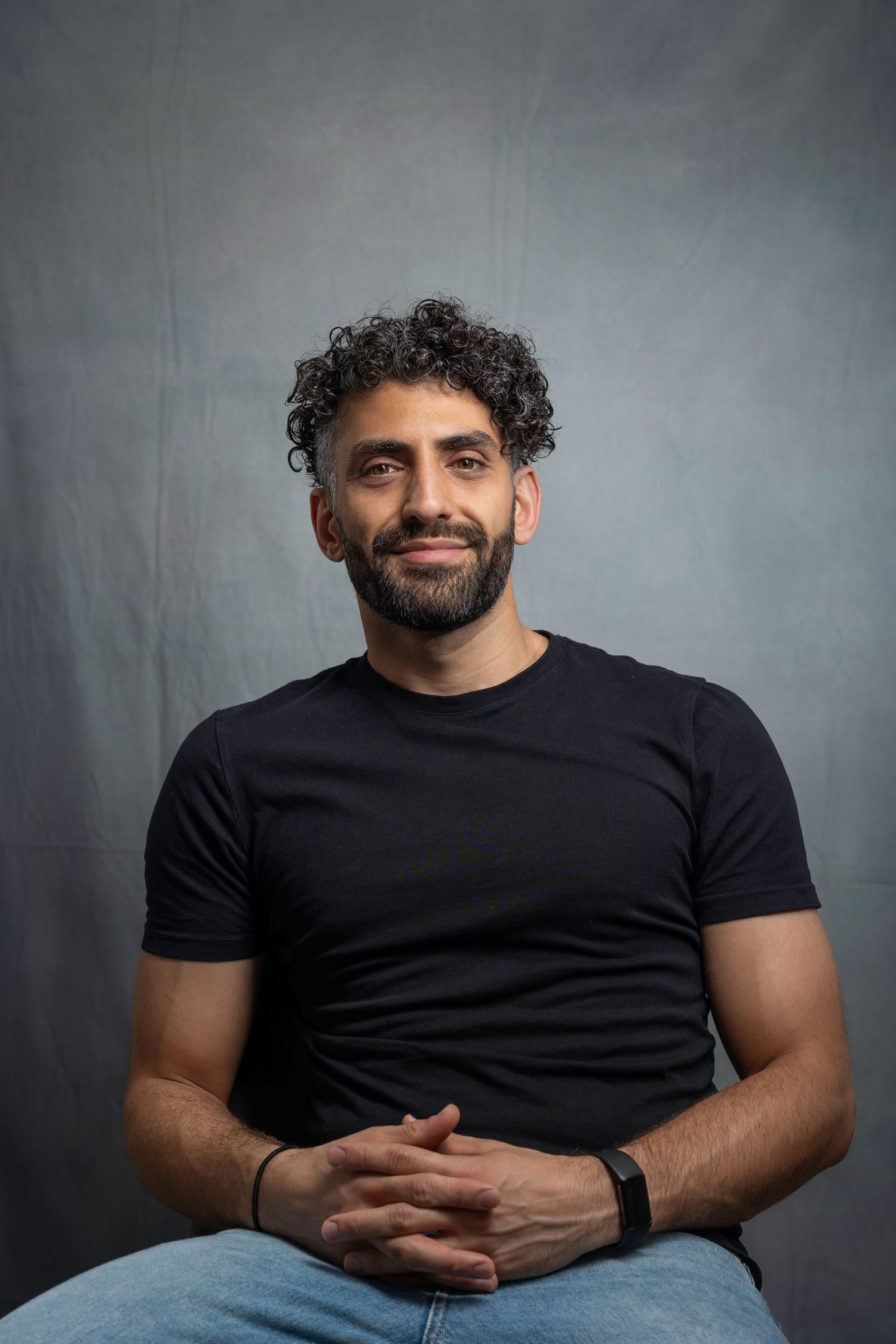 Smiling bearded man with curly hair in black t-shirt sitting with clasped hands in studio.