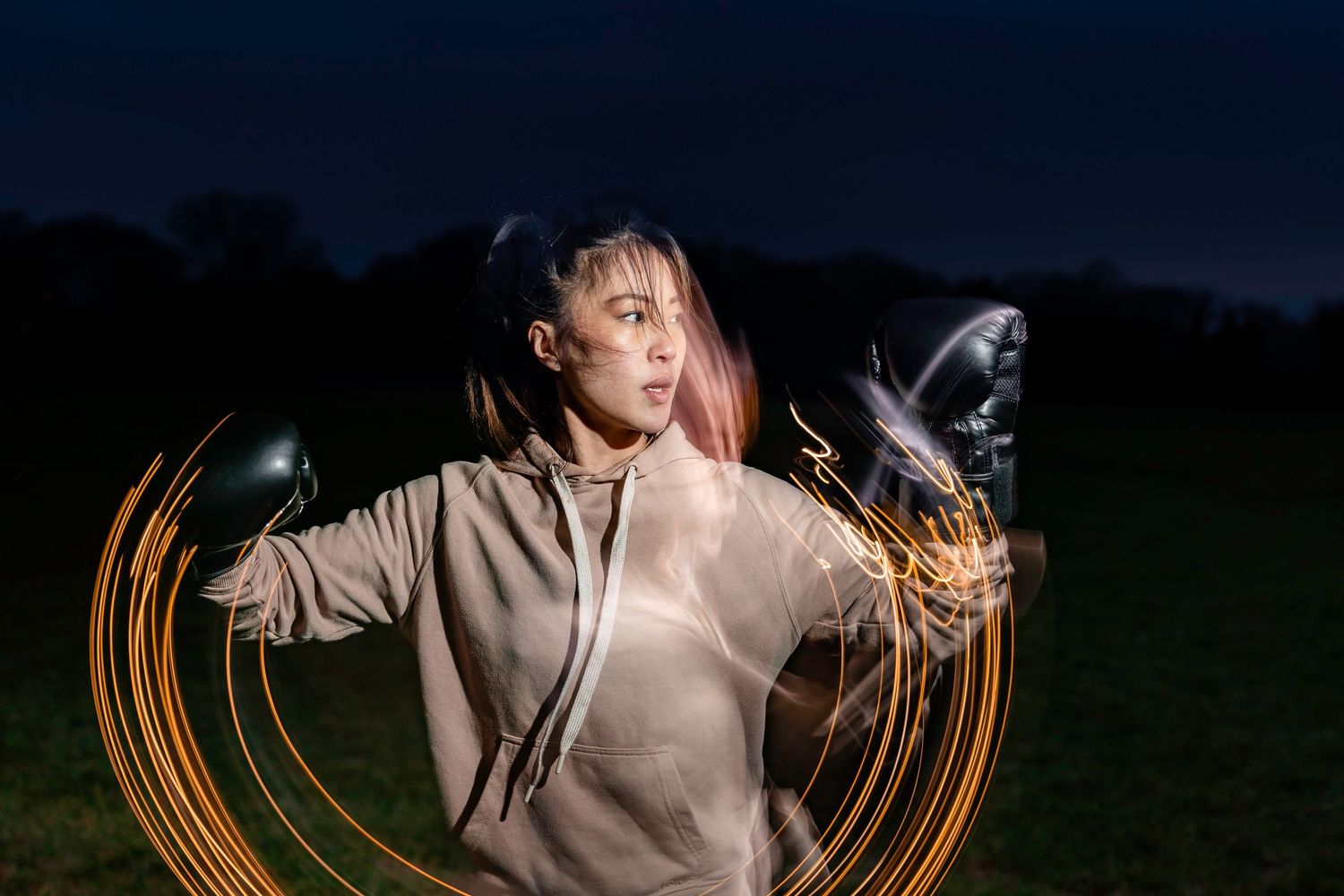 Woman in gray hoodie creating light painting art with glowing orange trails at night.