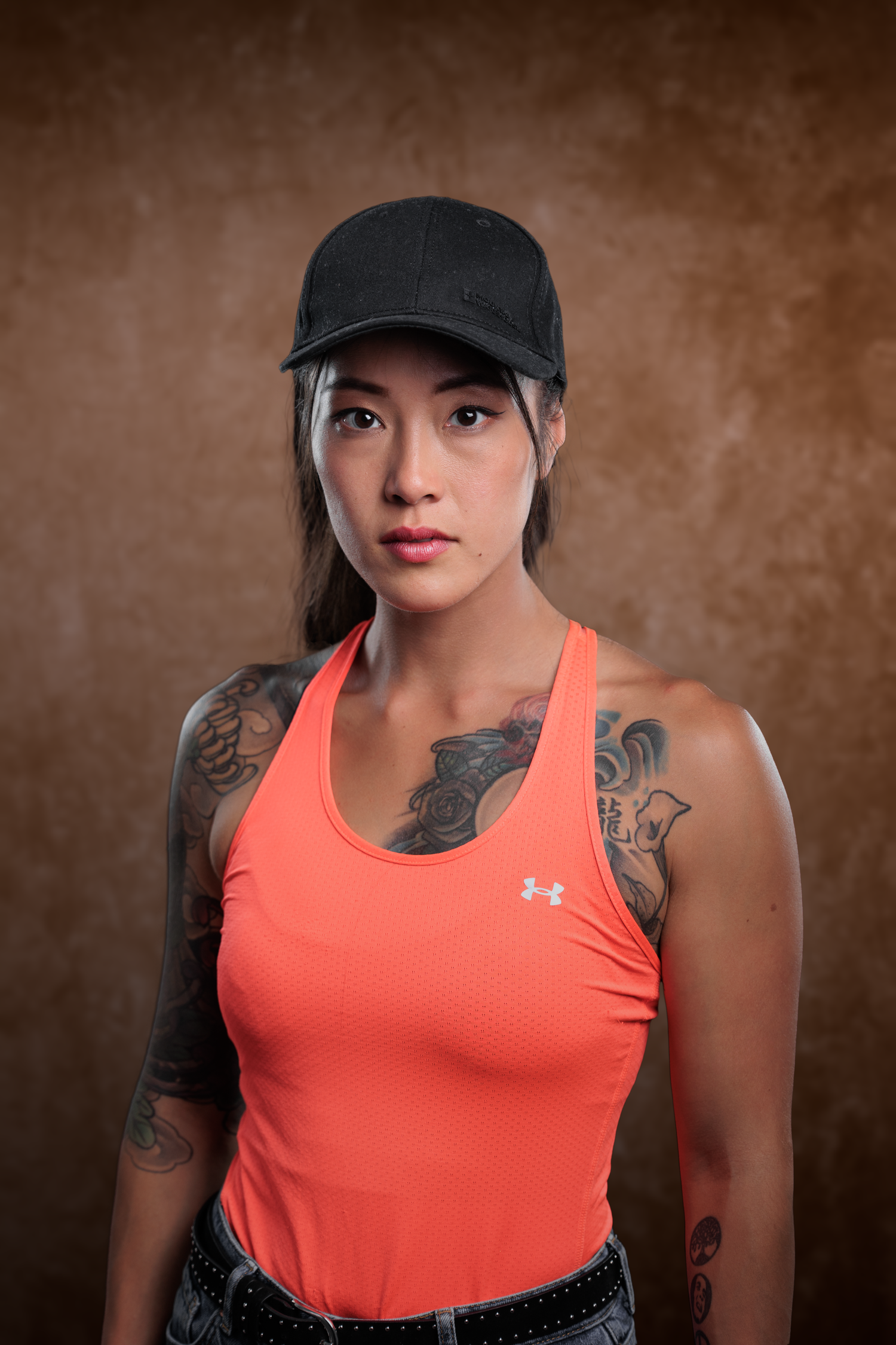 Tattooed woman in orange Under Armour tank top and black cap posing against brown studio backdrop.
