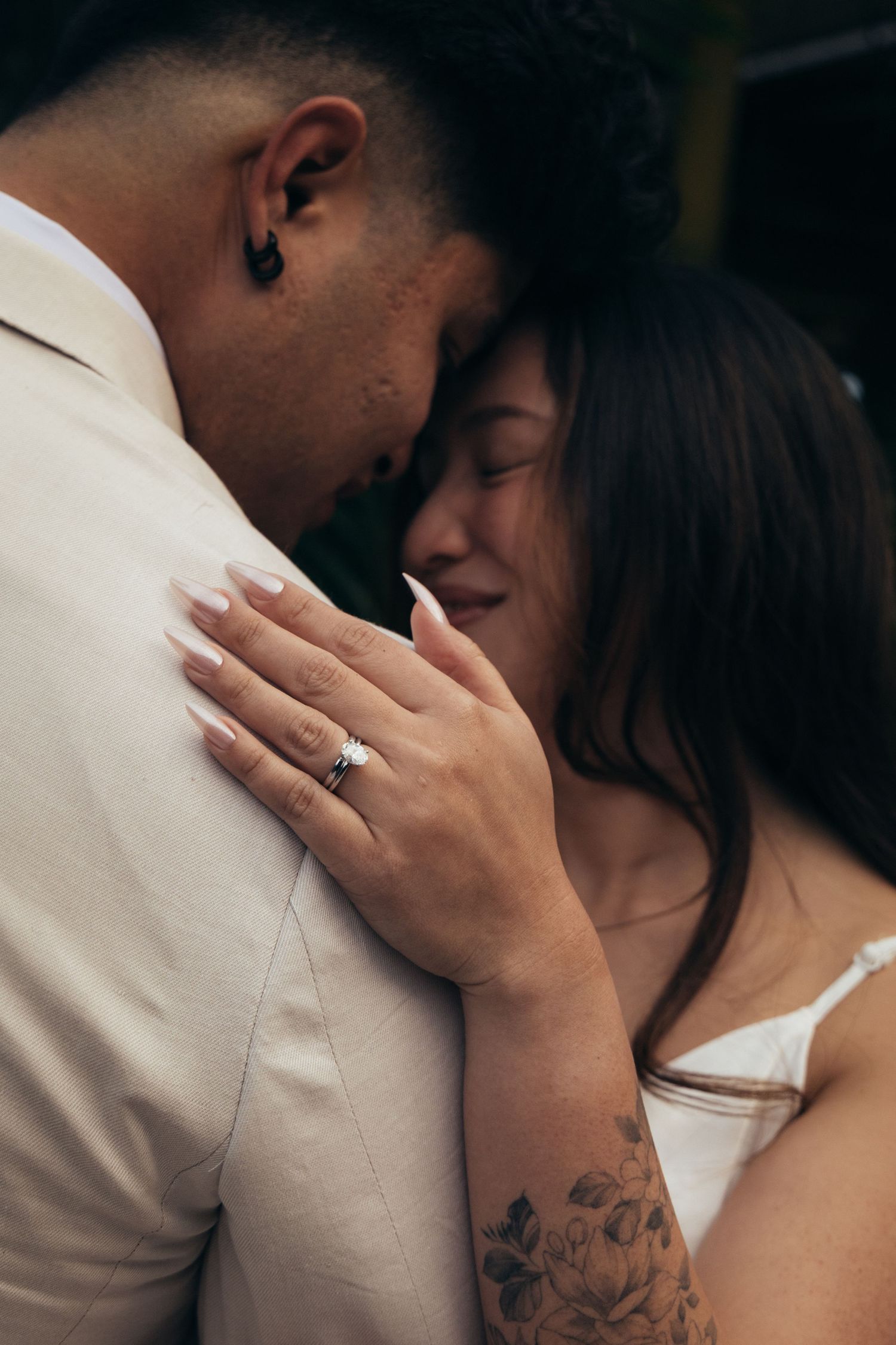 Couple sharing an intimate moment, woman displaying engagement ring and floral arm tattoo in a romantic close-up.