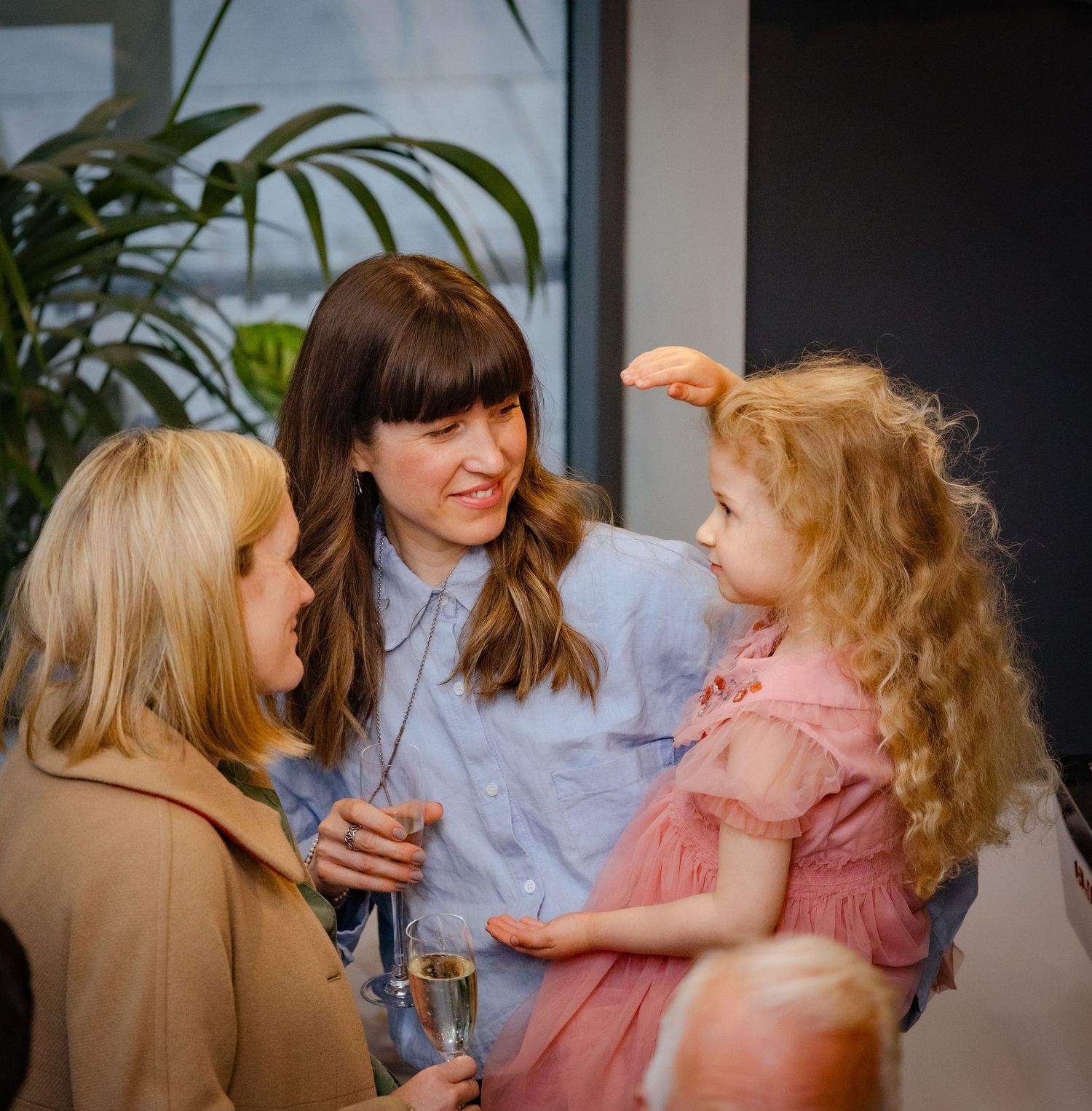 Two women interact with a young curly-haired girl in a pink dress at an indoor event.